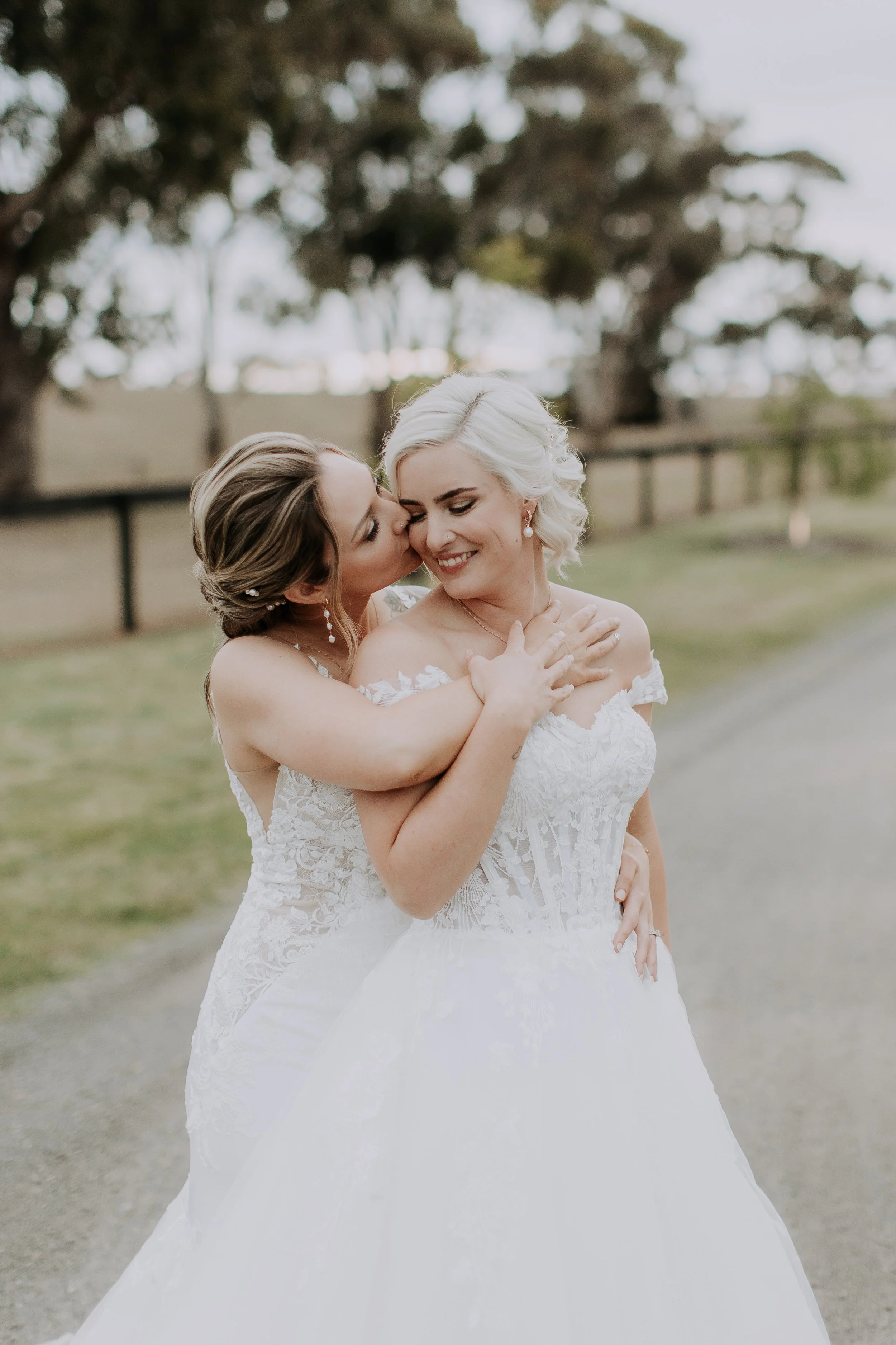 Two women in wedding dresses sharing a joyful moment, one kissing the other's cheek in an outdoor setting with trees in the background.