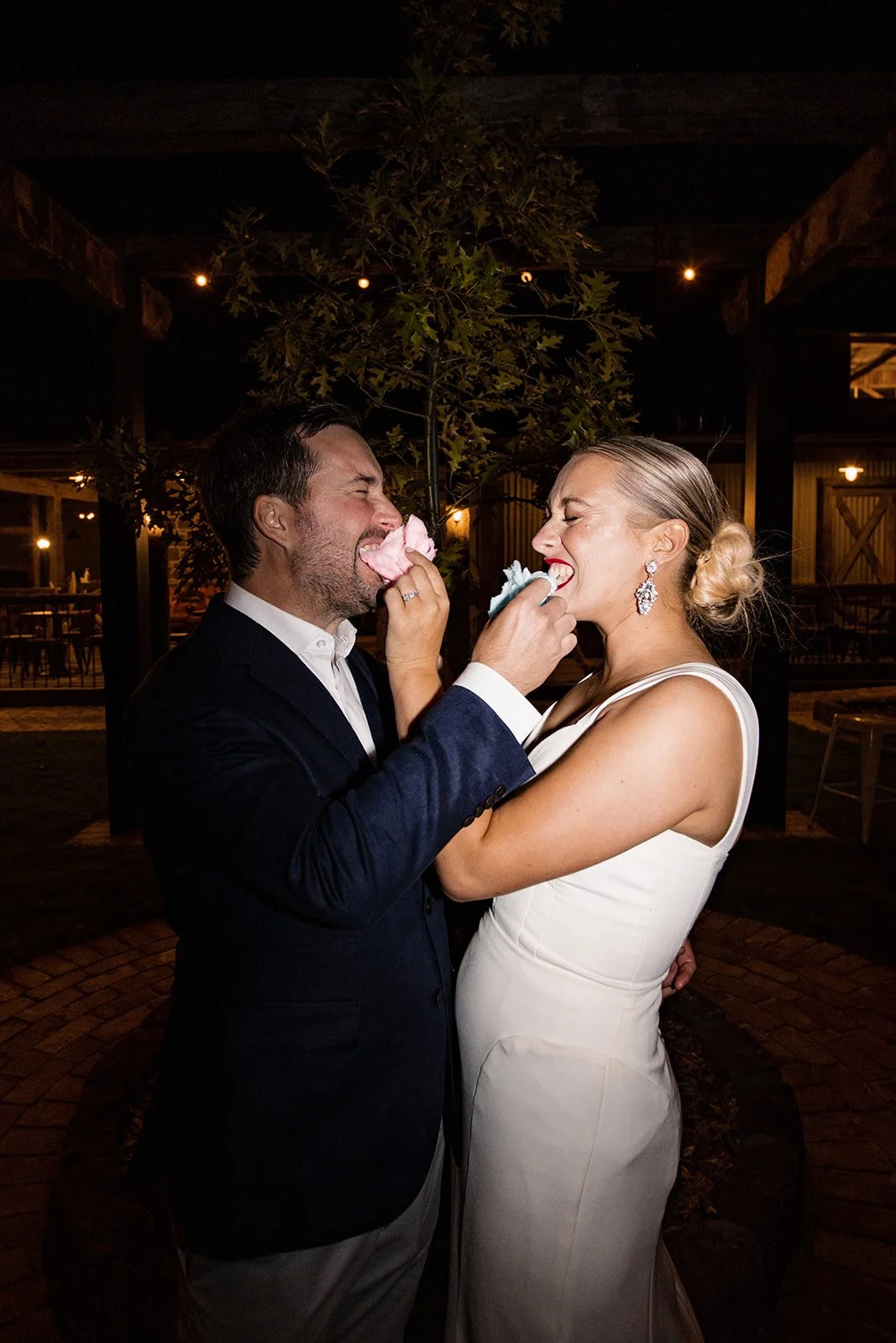 A bride and groom in an outdoor setting at night, playing with cotton candy snacks.