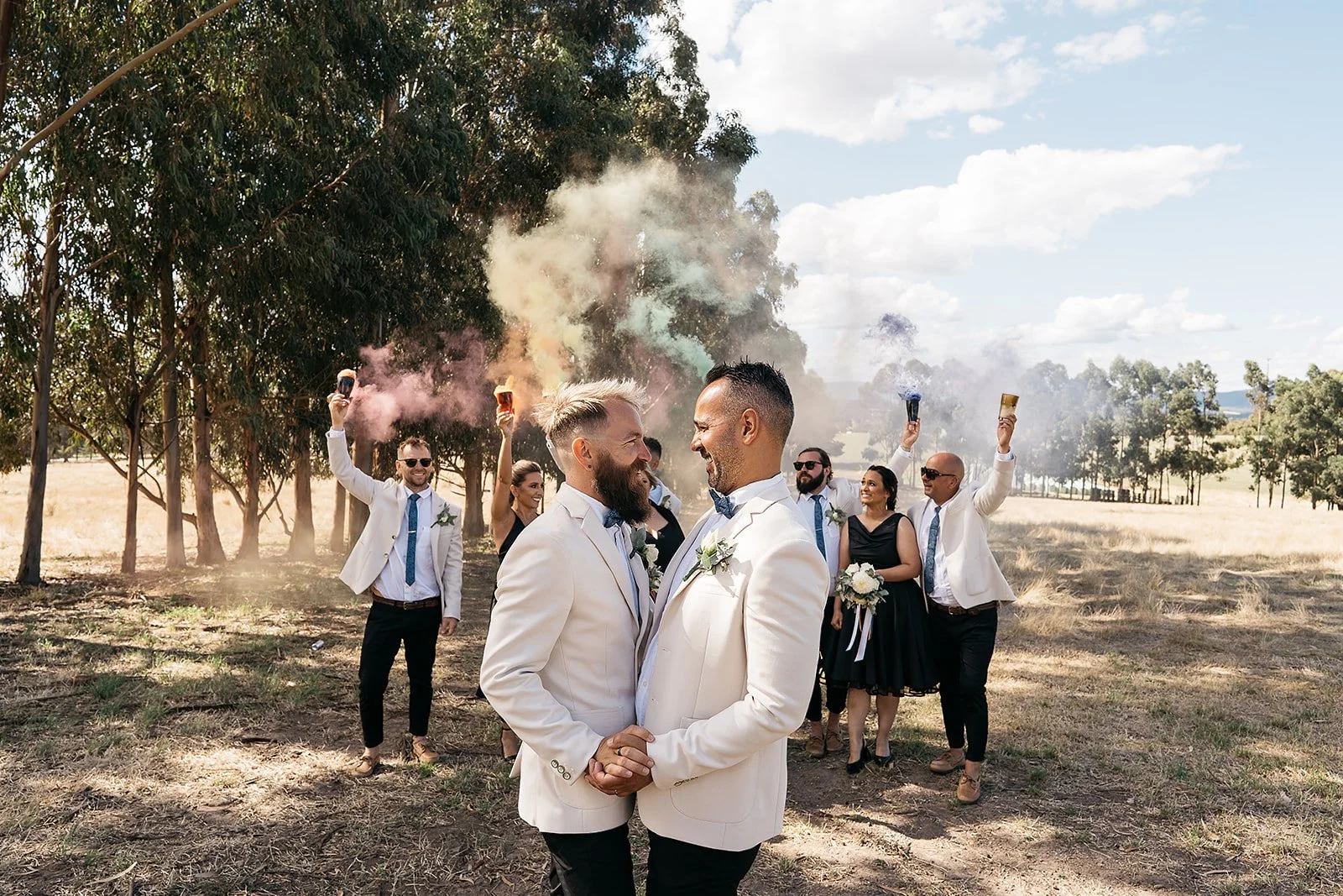 A same-sex couple in white suits holding hands and facing each other, surrounded by friends with colorful smoke flakes in an outdoor field.
