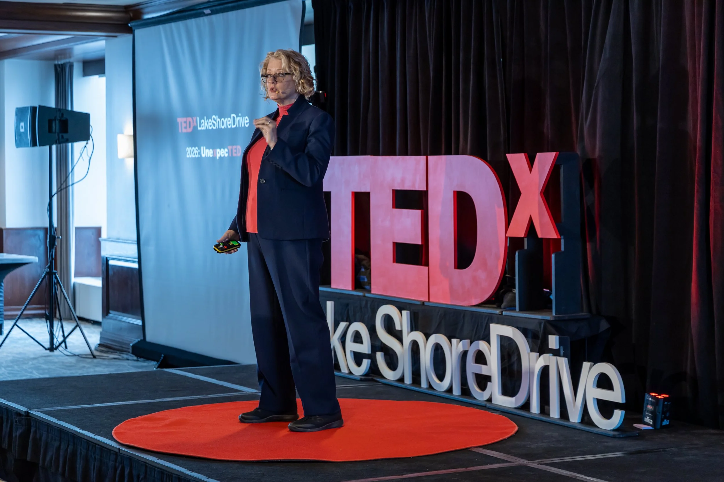 A woman speaking on stage at a TEDx event, standing on a circular red carpet with a large TEDx sign behind her. She is dressed in a suit and glasses, holding a remote control device in her hand.