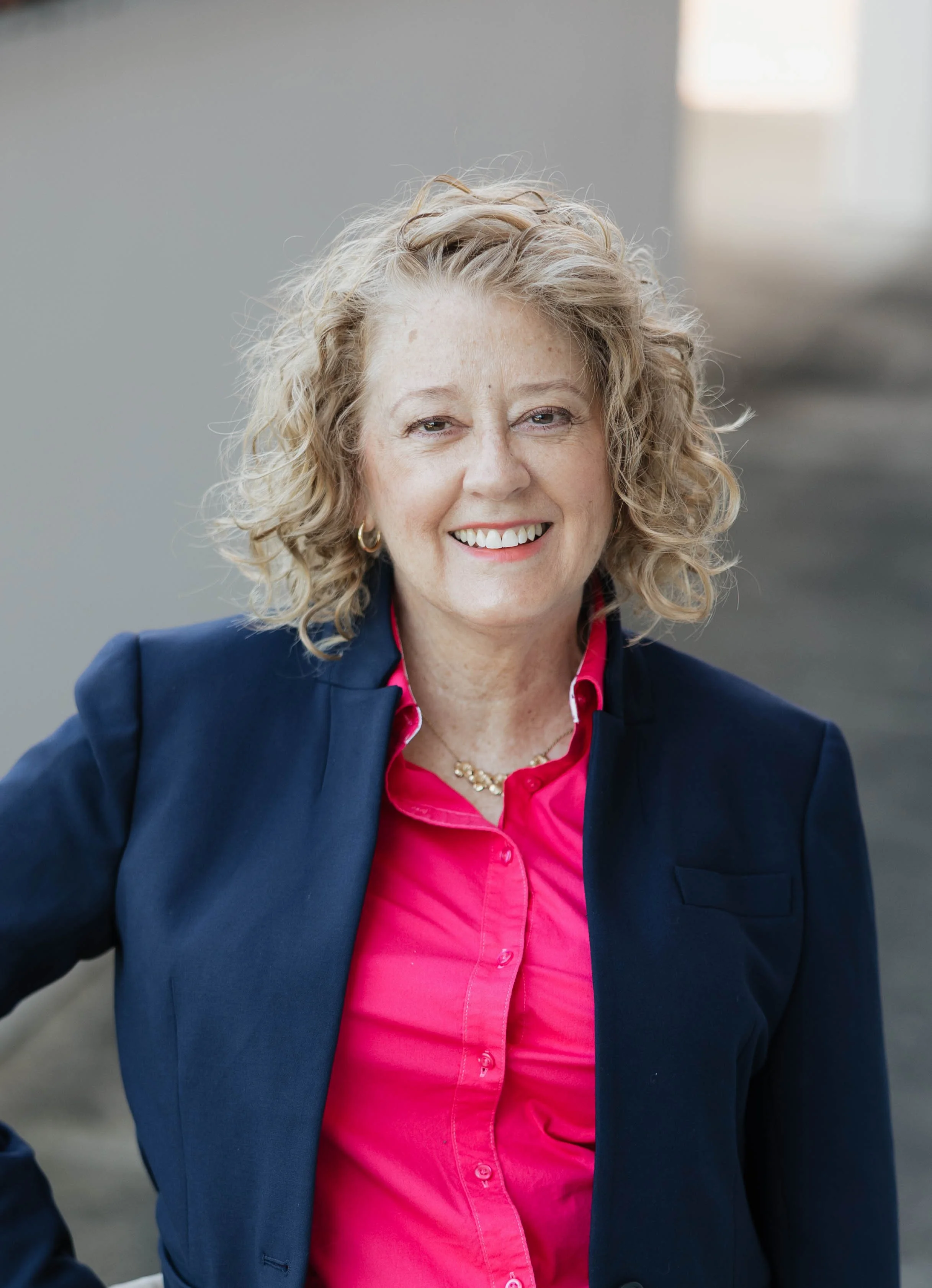 A smiling middle-aged woman with curly blonde hair, wearing a navy blazer over a bright pink shirt, in a well-lit indoor space.