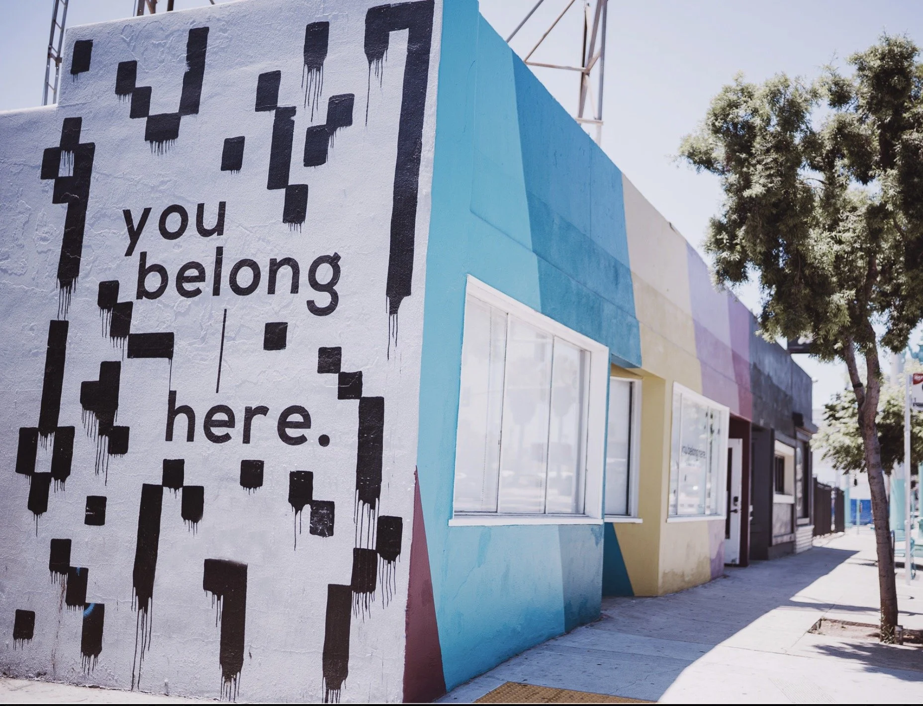 Colorful mural on a building wall featuring the words "you belong here" with black pixelated shapes hanging from the top.