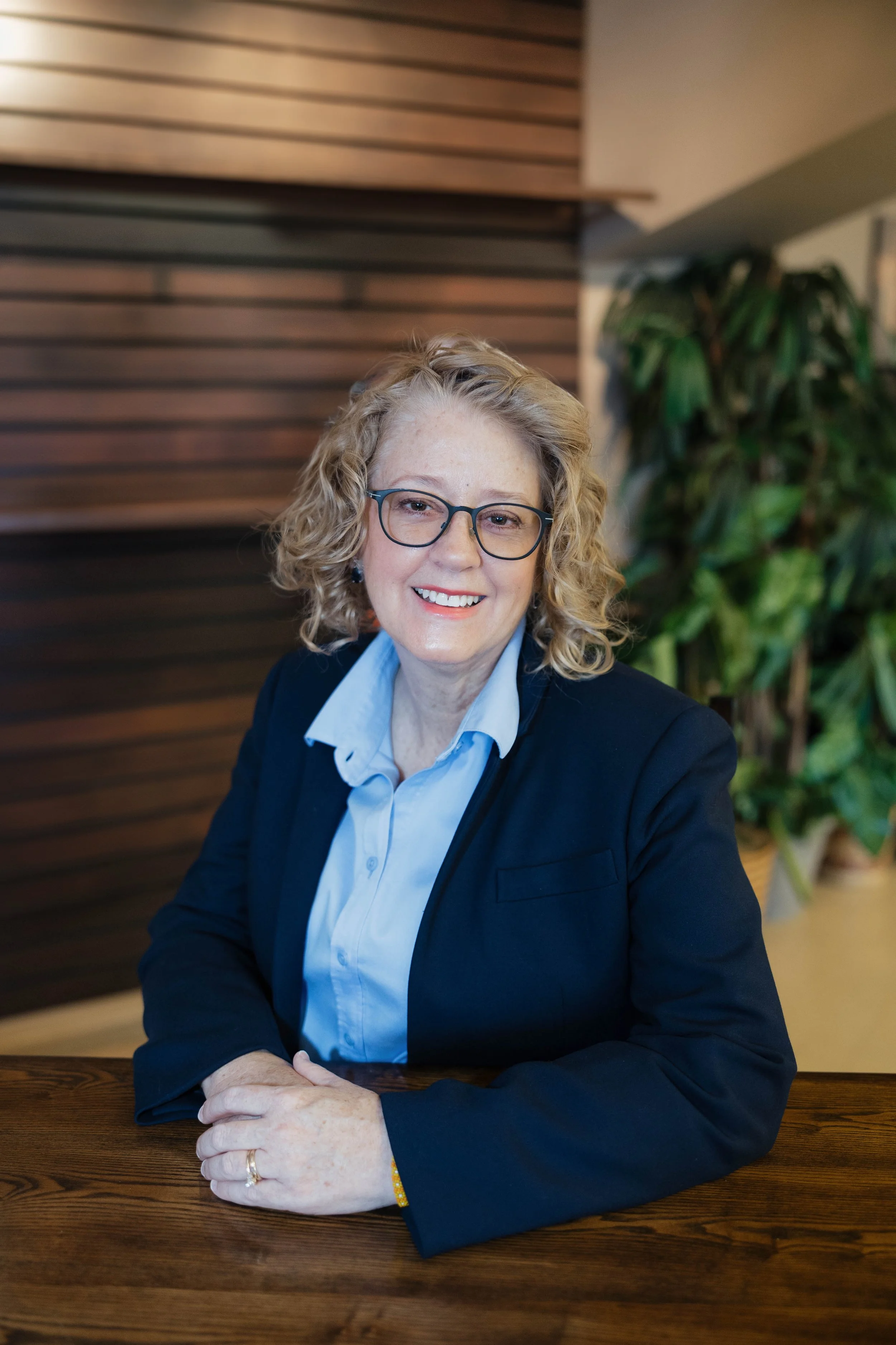 A woman with curly blonde hair, glasses, and a friendly smile sitting at a wooden table, wearing a navy blazer and a light blue shirt, in a modern interior with wooden wall paneling and a green plant in the background.