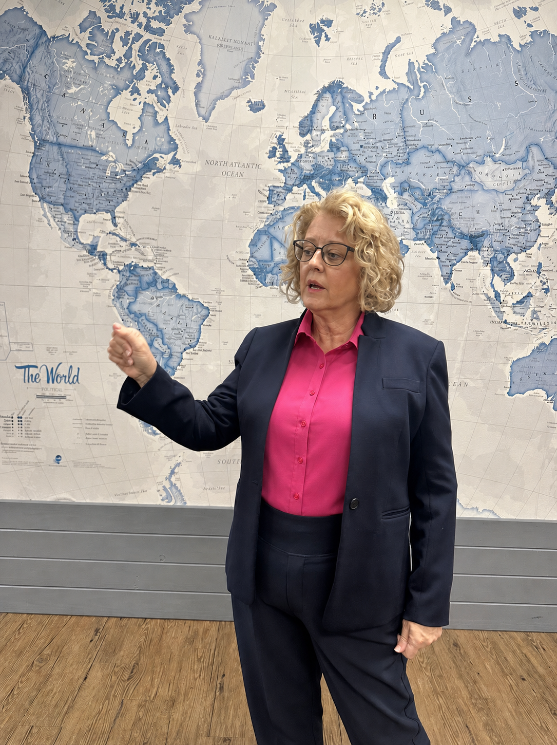 A woman with curly blonde hair, glasses, wearing a navy blazer and pink blouse, pointing at a world map behind her.