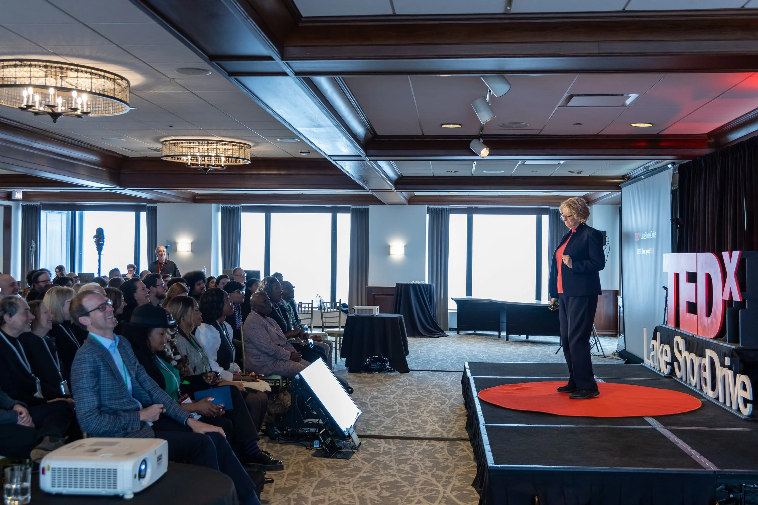 A woman stands on a stage giving a TED talk presentation in a conference room filled with an audience.