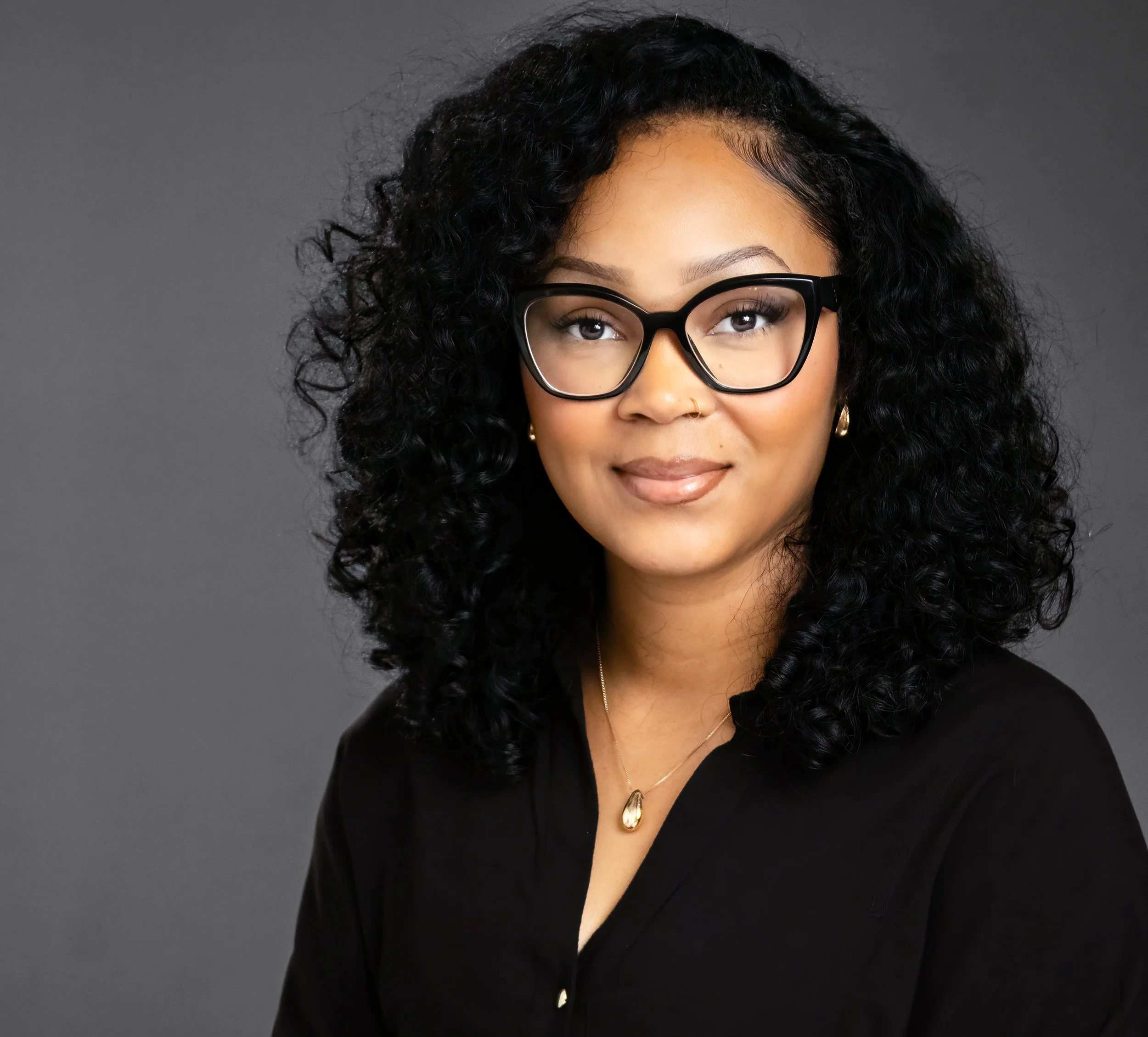Close-up of a woman with curly black hair, wearing glasses, gold hoop earrings, a gold necklace with a teardrop pendant, and a black blazer, against a plain gray background.