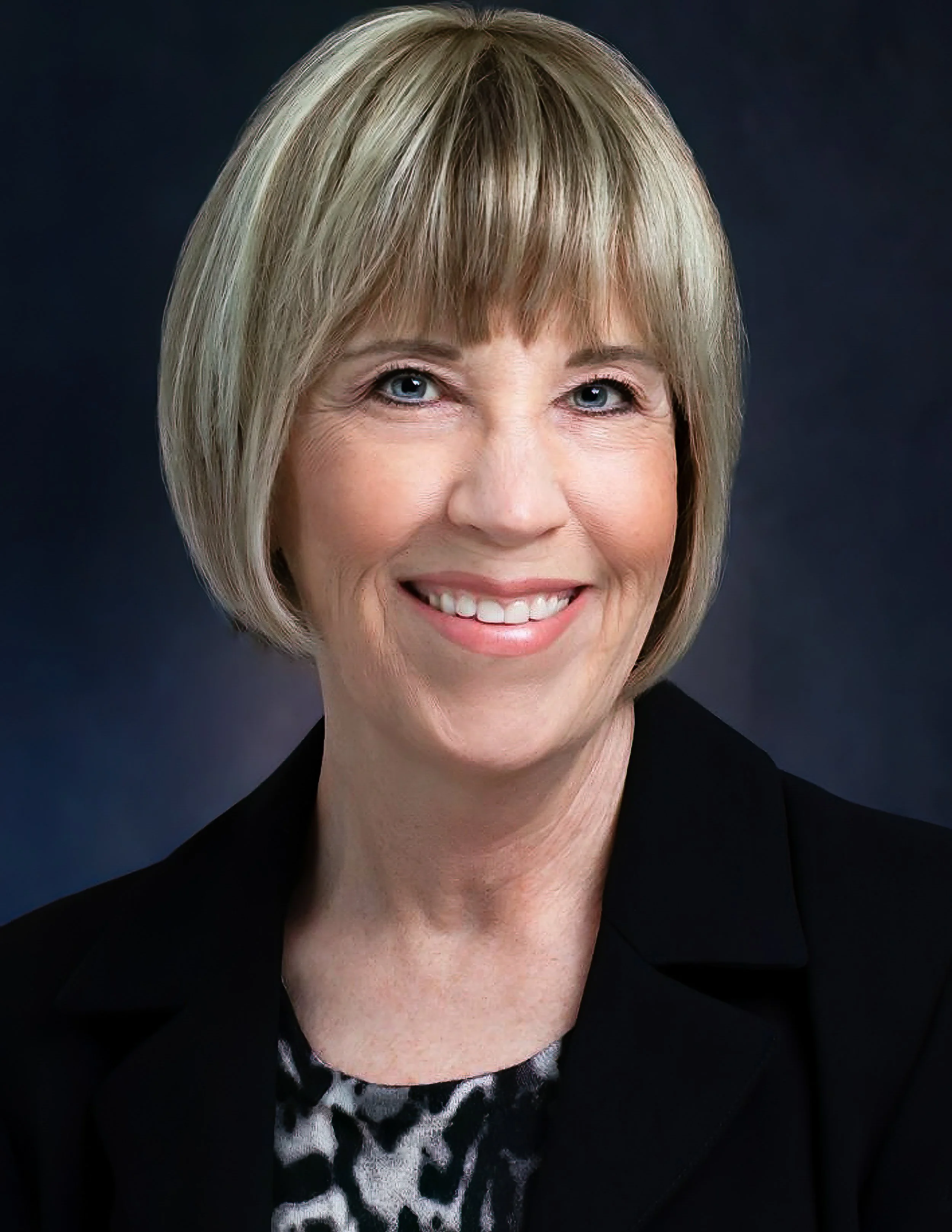 Close-up of a smiling woman with short blonde hair, wearing a black blazer and a leopard print blouse, against a dark background.