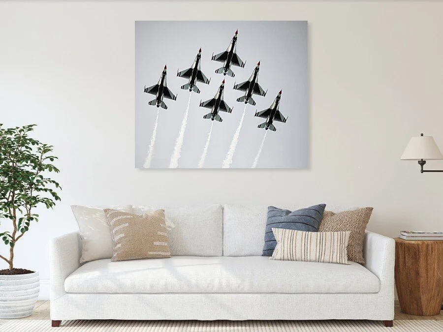 A painting of seven fighter jets flying in formation, leaving white exhaust trails, hanging on a living room wall above a white sofa with several pillows.
