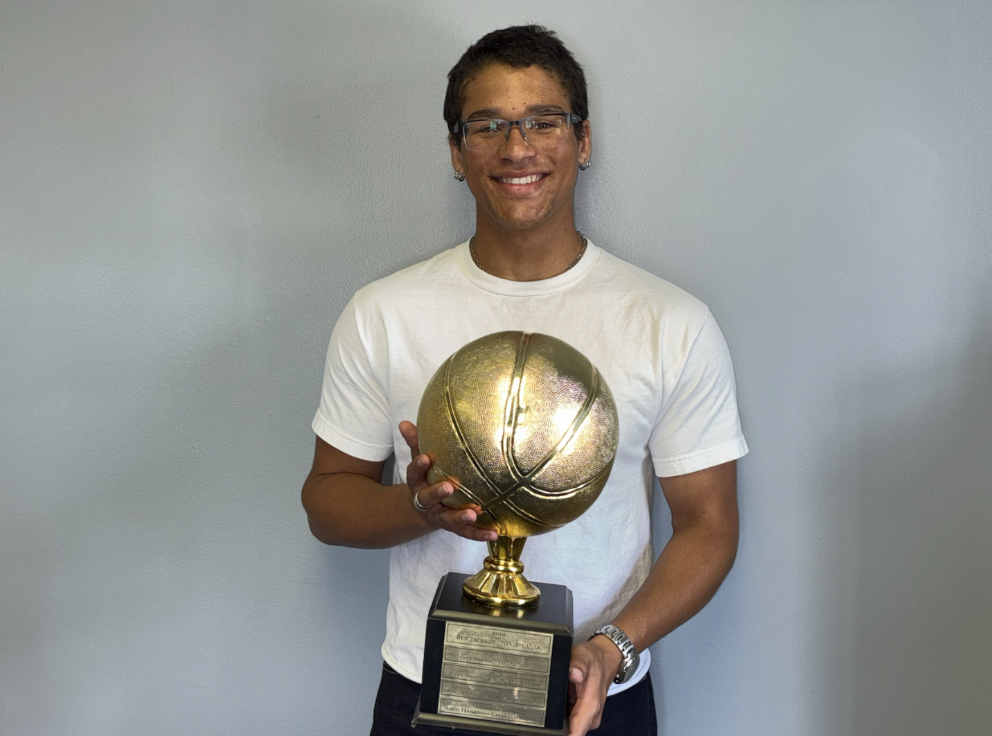 A smiling young man with glasses and earrings, wearing a white t-shirt, holding a gold-colored basketball trophy with a plaque at the base, standing against a plain gray wall.