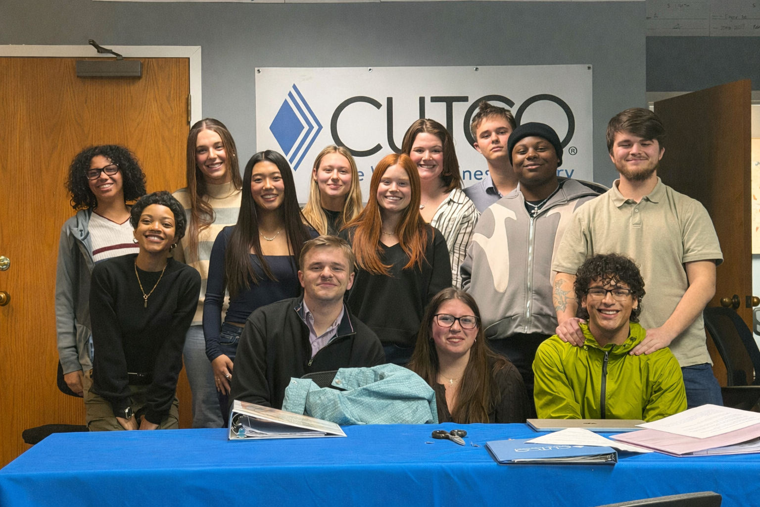 Group of young people posing together at a meeting or event, standing behind a table with papers and folders, with a sign that reads 'CITGO' in the background.