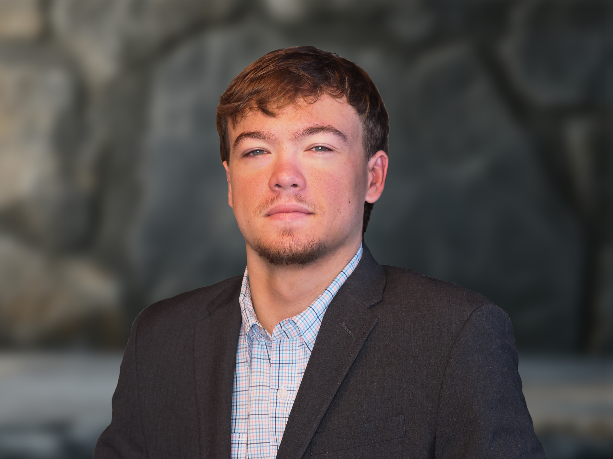 A young man with brown hair and a goatee wearing a dark suit jacket over a plaid shirt, standing outdoors in front of a blurred rocky background.