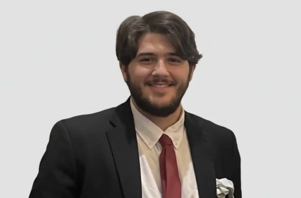 A young man with dark hair and a beard, wearing a black blazer, white shirt, and red tie, smiling at the camera against a plain white background.