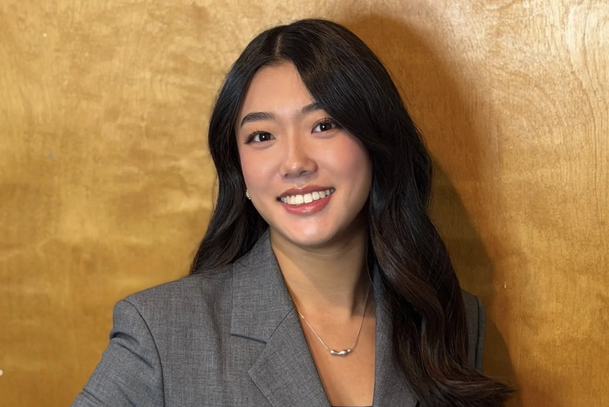 A young woman with long, wavy black hair, wearing a gray blazer and a silver necklace, smiling in front of a wooden background.