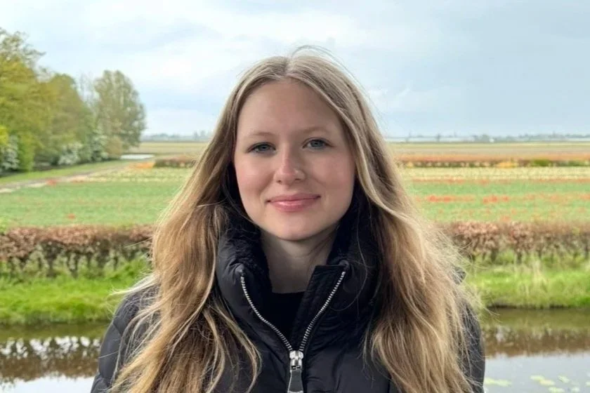 A young woman with long blonde hair and a black jacket standing outdoors in front of a field and a pond on a cloudy day.