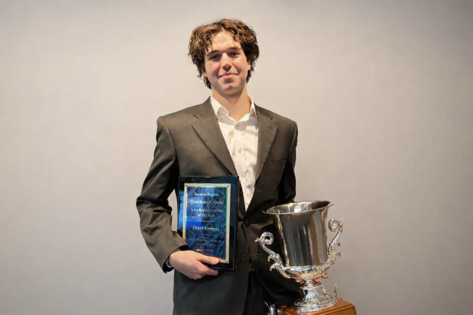 A young man with curly brown hair dressed in a gray suit and white shirt standing against a plain light-colored background, holding a framed certificate or award in his right hand and a large silver trophy with ornate handles in his left hand.