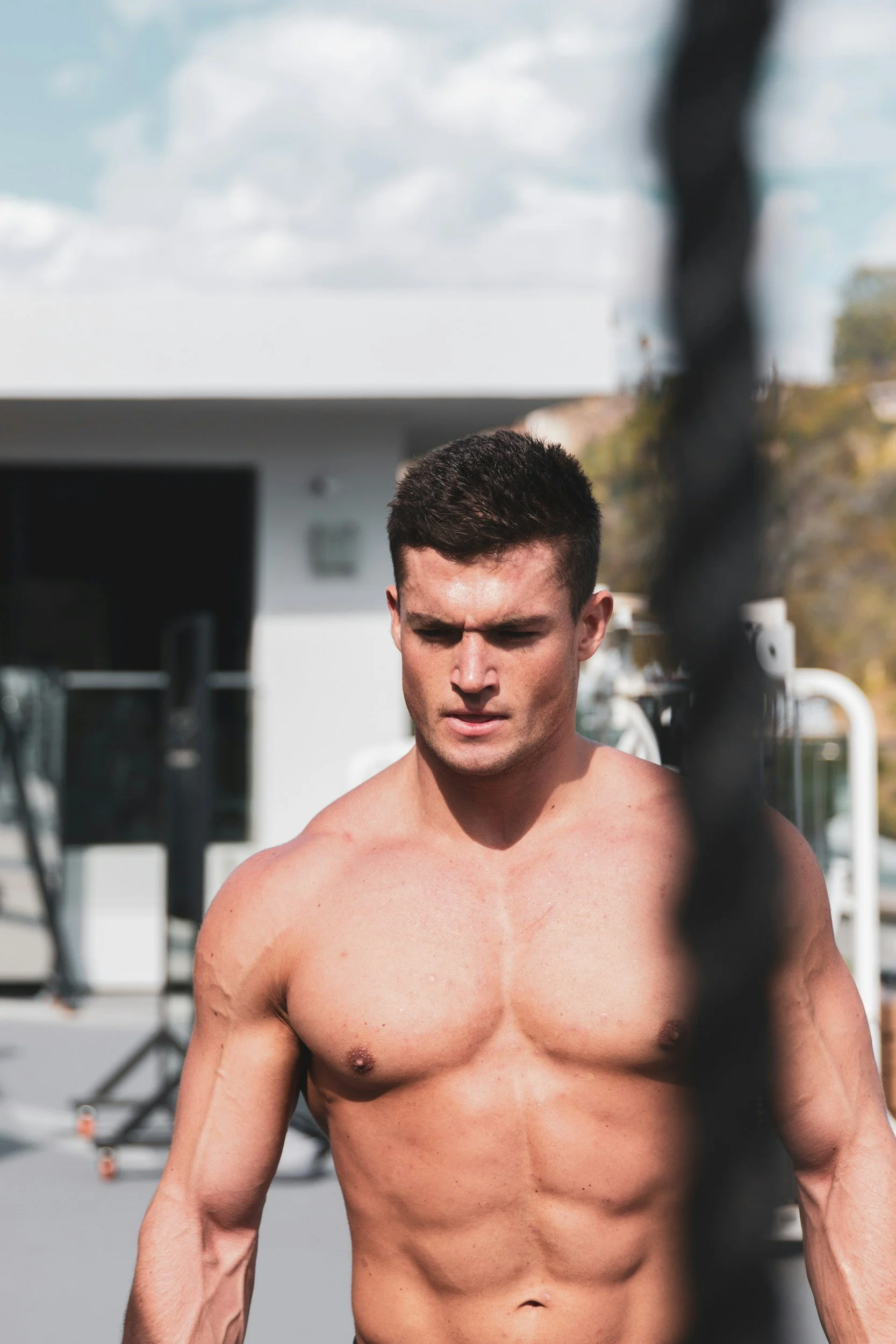 A shirtless man with short dark hair and muscular build looking downward outdoors, with a blurred background of a building, trees, and clouds.