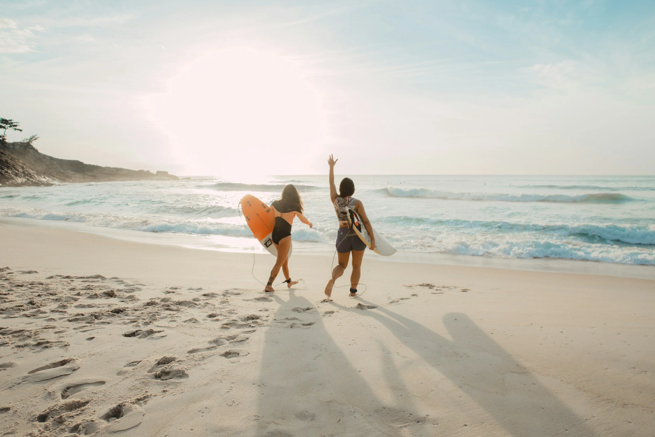 Two people holding surfboards walking away from the camera on a sandy beach toward the ocean during sunset. One person is raising their hand, and the scene features waves and a partly cloudy sky.