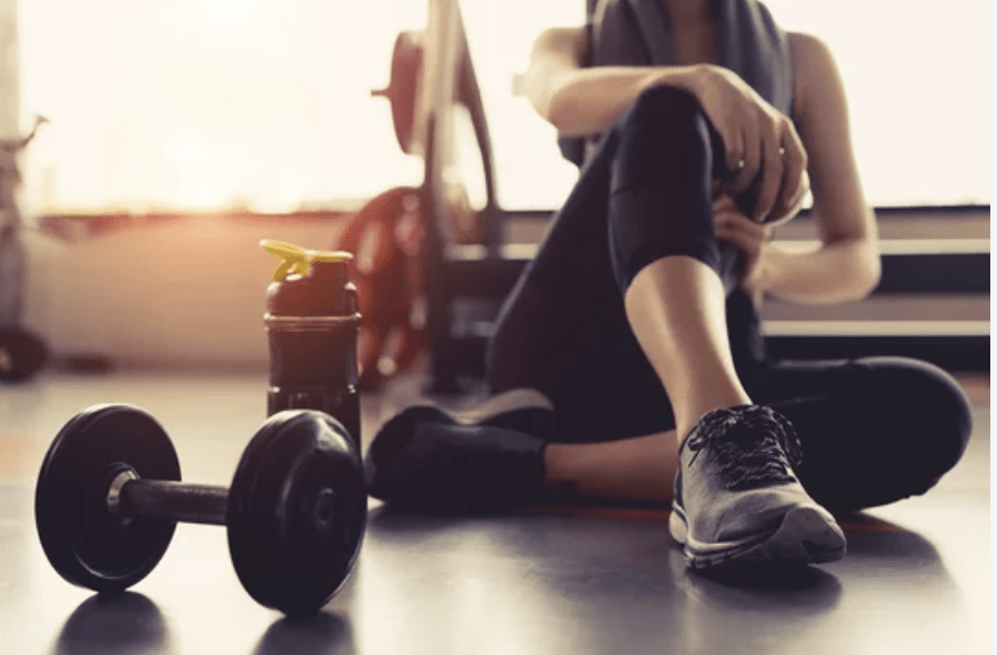 A woman sitting on the gym floor next to a water bottle and a dumbbell, dressed in workout attire.