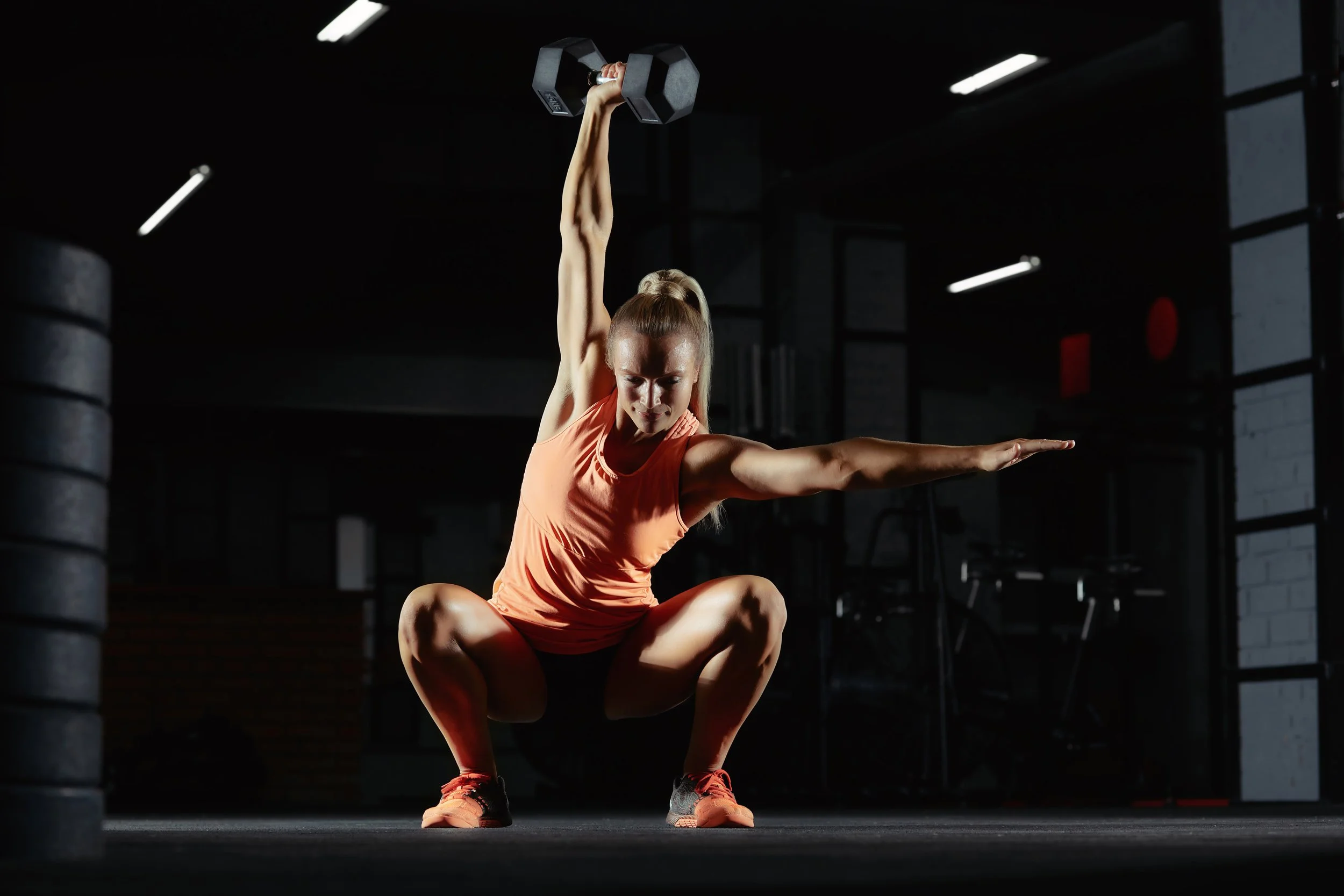 A woman in sportswear performing a squat exercise while holding a dumbbell overhead in a dimly lit gym.