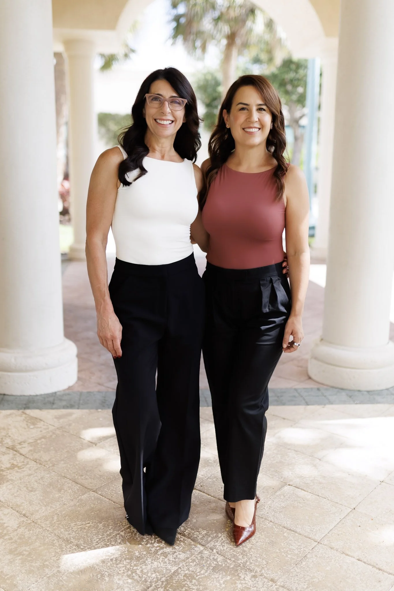 Two women standing together outdoors in front of white columns and trees, smiling at the camera. One woman has black hair, glasses, and is wearing a white sleeveless top with black pants. The other woman has brown hair and is wearing a sleeveless pink top with black pants and brown shoes.
