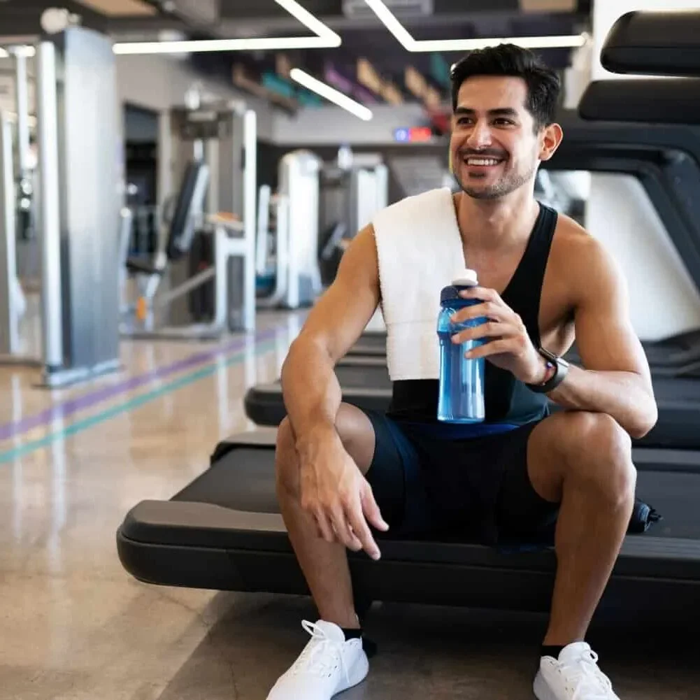 A smiling man sitting on a treadmill at the gym, holding a blue water bottle with a towel draped over his shoulder.