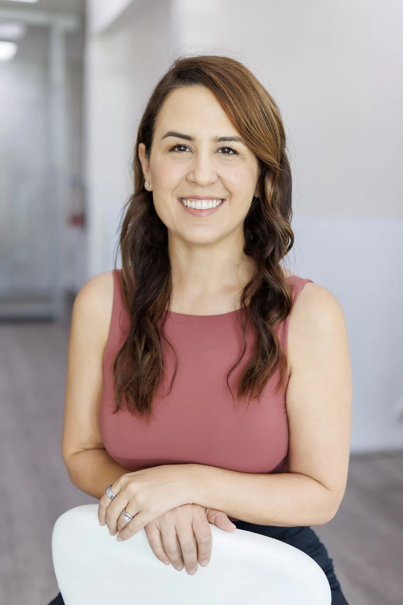 A woman with long, wavy brown hair and a broad smile, wearing a sleeveless pink top and rings on her fingers, sitting in a modern, bright office space with gray walls.