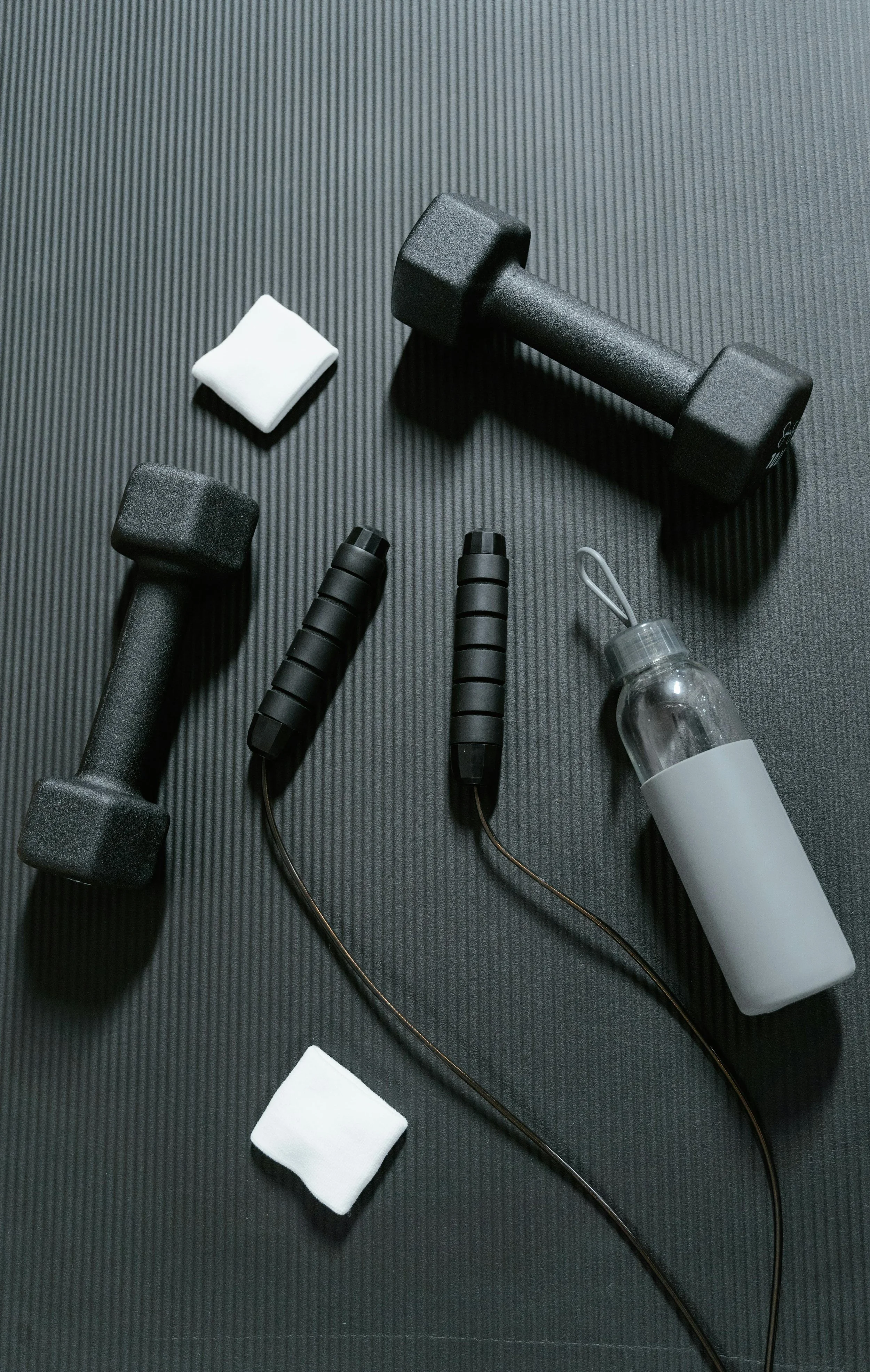 Dumbbells, jump rope, water bottle, and cleaning cloth on black workout mat.
