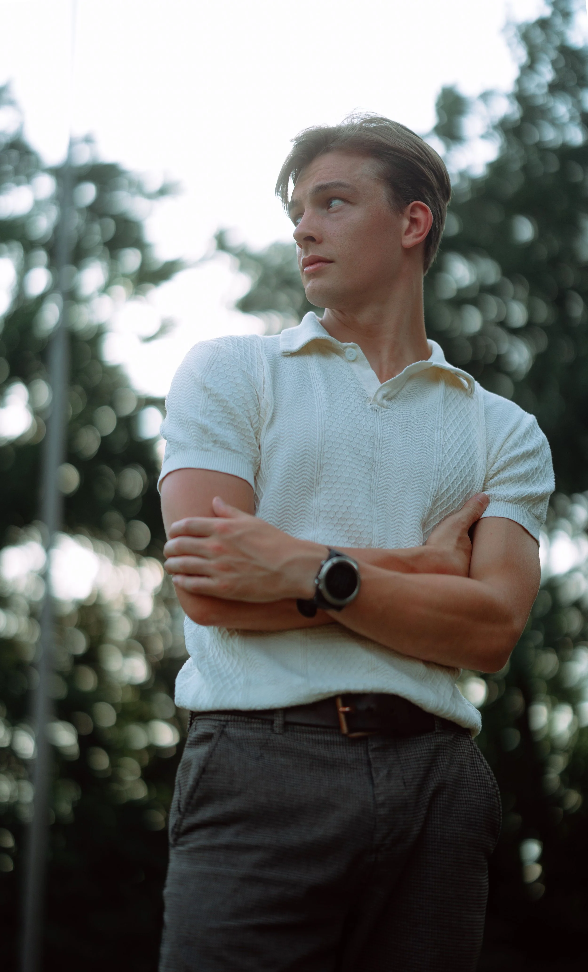 A young man with light skin and brown hair standing outdoors with trees in the background. He is wearing a white textured polo shirt, gray plaid pants, and a black wristwatch. He has his arms crossed and is looking to the side with a thoughtful expression.