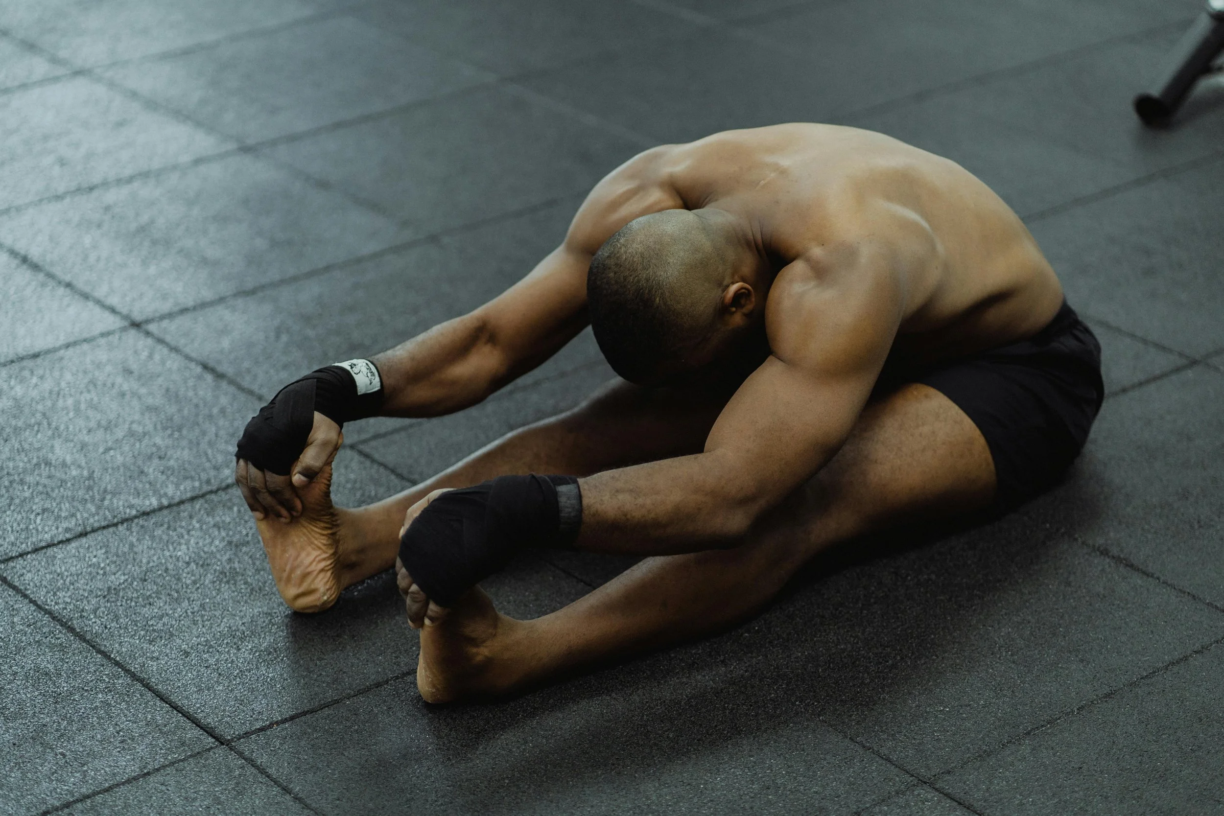 A shirtless man wearing black shorts and black wrist wraps is sitting on the floor, stretching by reaching towards his toes with both hands, with his head bent down.