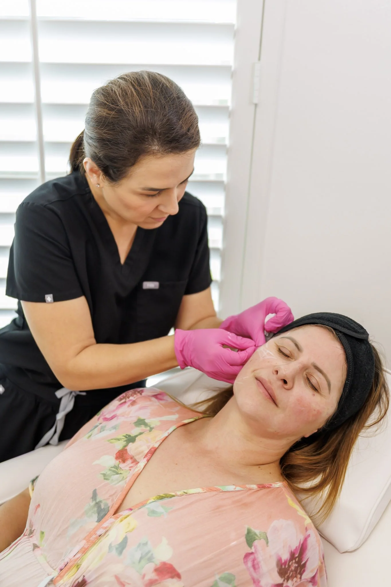 A woman is lying on a medical bed with her head resting on a pillow, wearing a floral shirt. A medical professional, dressed in black scrubs with pink gloves, is gently administering an injection or performing a treatment on her face. The setting appears to be a medical or dermatology clinic with white walls and window blinds in the background.