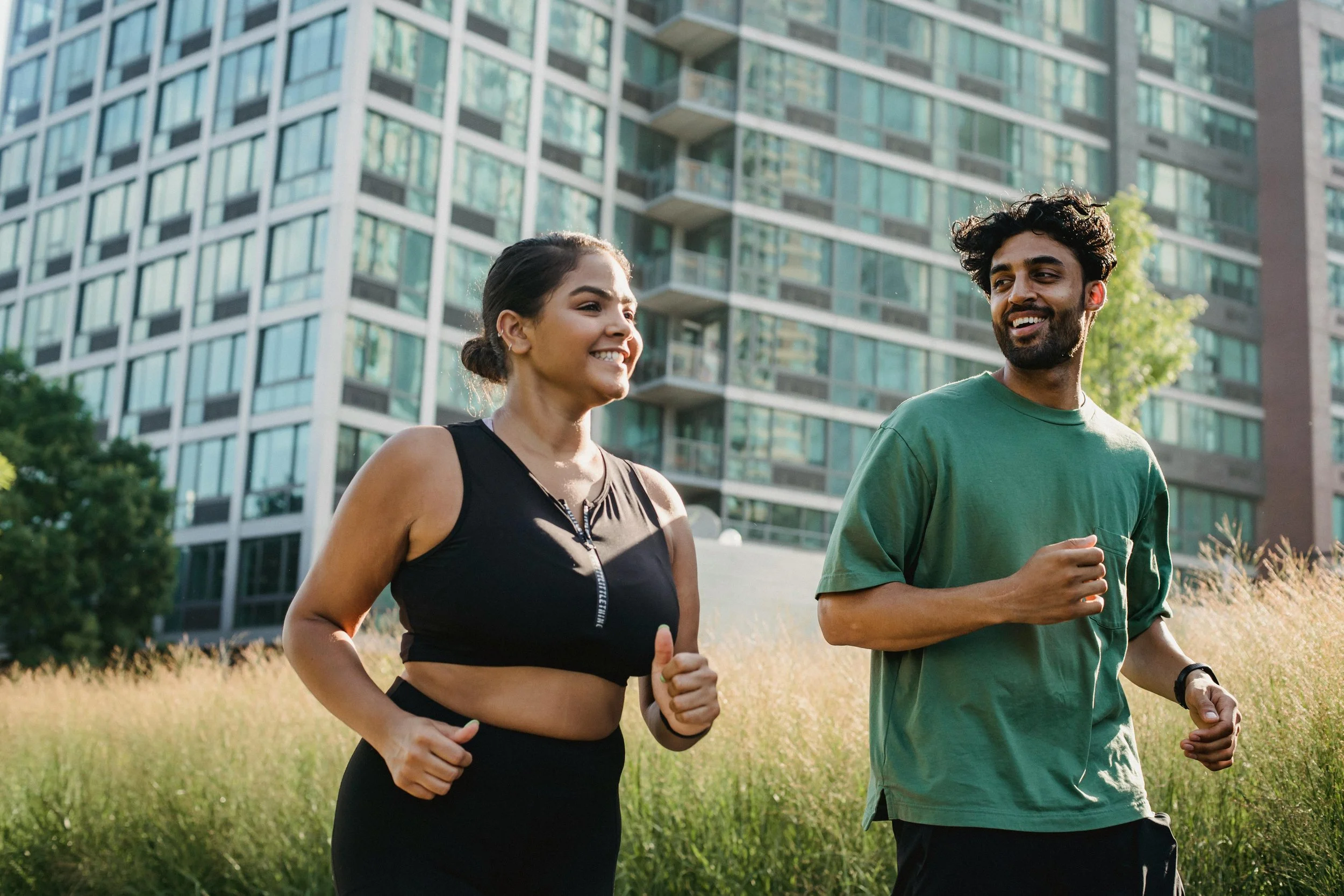 A woman and a man running outdoors in a grassy area with tall buildings in the background, smiling and enjoying exercise.