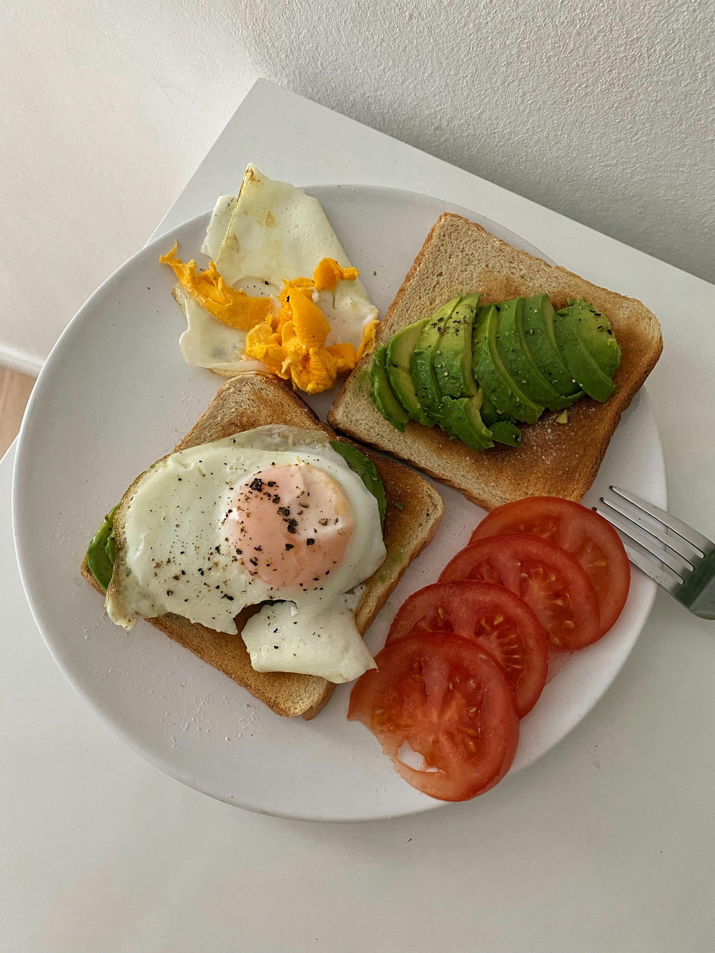 Plate with two slices of toast, one topped with sliced avocado, and the other with a fried egg. Accompanied by sliced tomatoes, a few pieces of scrambled eggs, and a couple of egg whites.