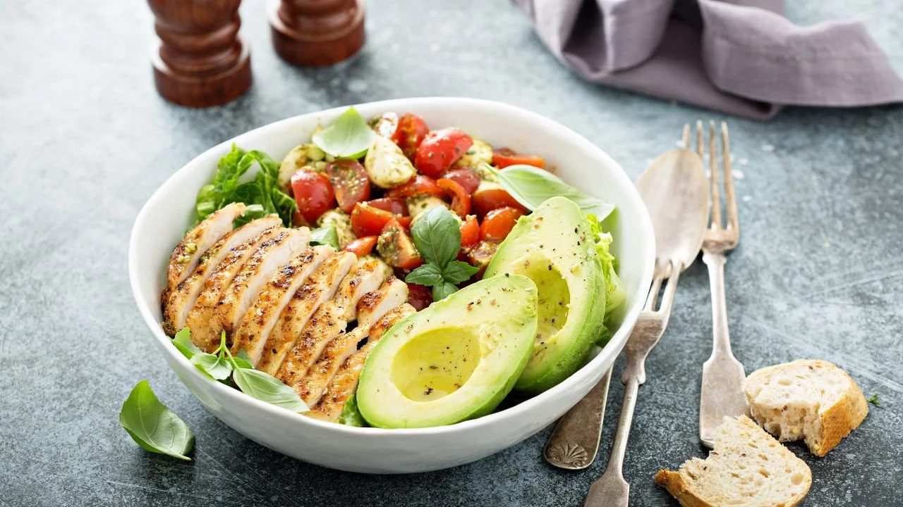 A white bowl filled with sliced grilled chicken, avocado halves, and a fresh tomato salad on a gray surface, with a fork, knife, and piece of bread nearby.