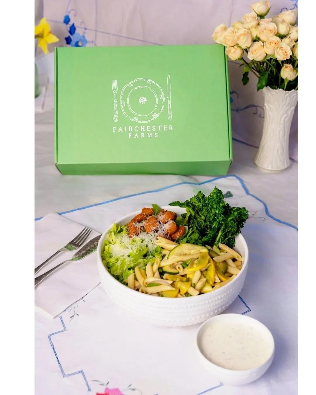 A salad in a white bowl with lettuce, broccoli, pasta, and shredded cheese, accompanied by a small cup of creamy dressing, placed on a table with a white tablecloth and silverware. In the background, there is a decorative box from Fairchester Farms, a white vase with pink roses, and some blue and yellow decorations.