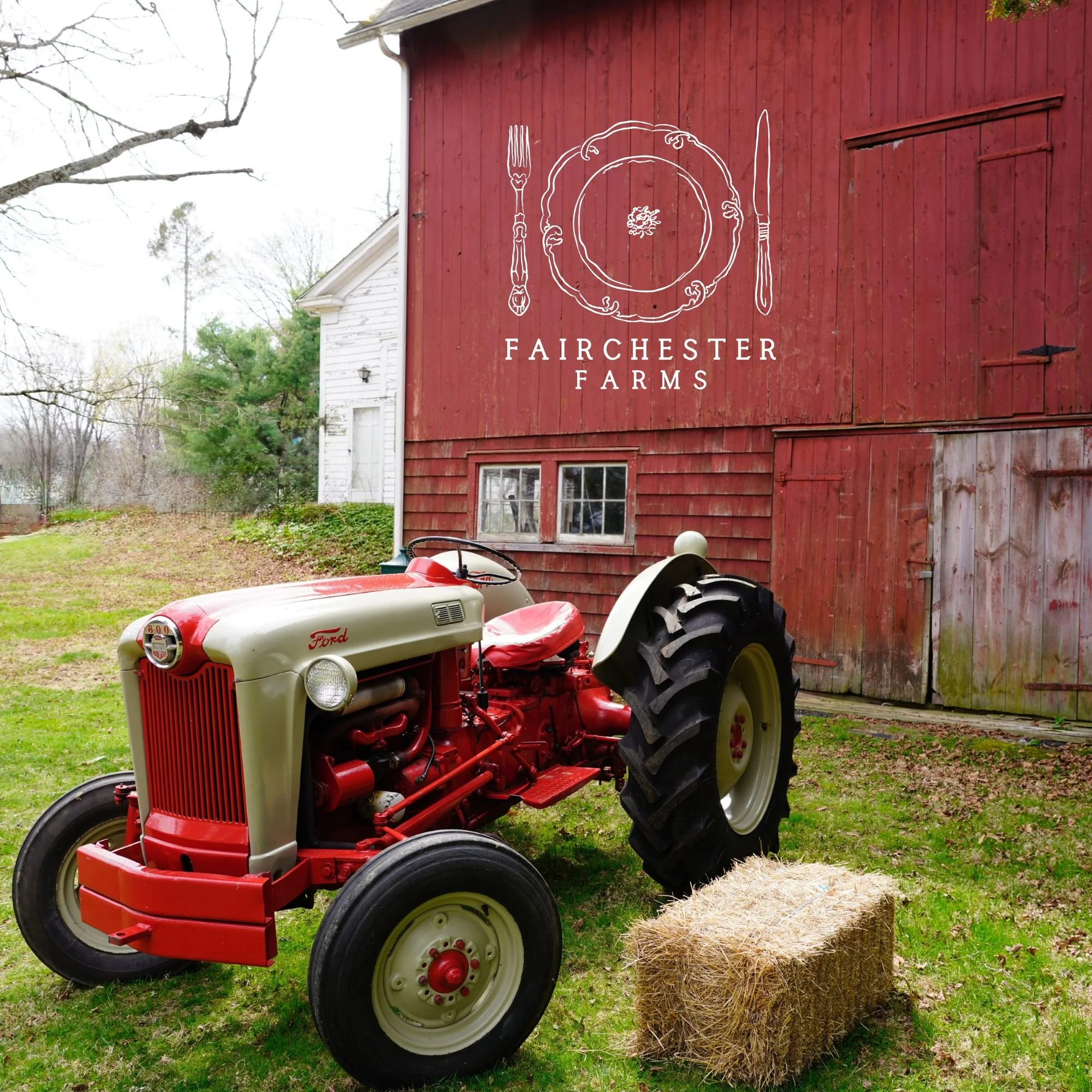 A vintage Ford tractor parked on a grassy area in front of a red barn with a "Fairchester Farms" logo. There is a hay bale on the ground nearby.