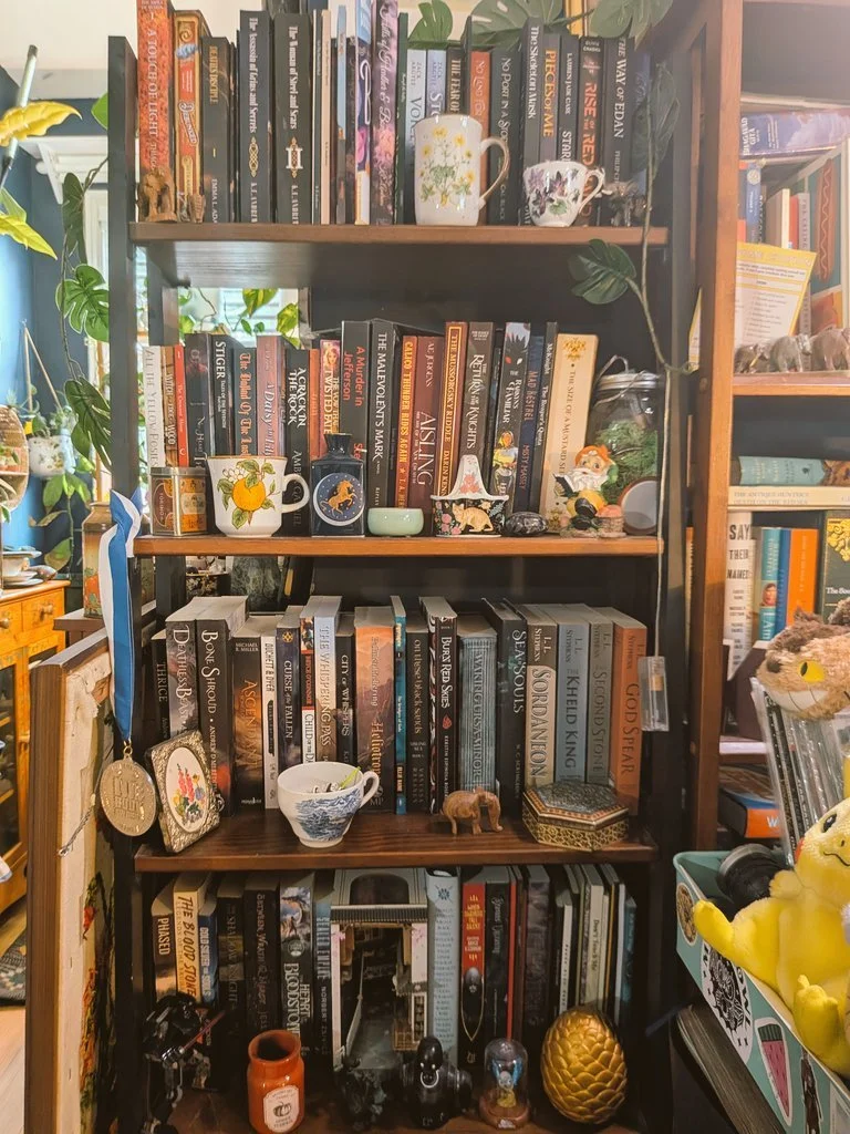 Wood bookshelf filled with travel, fantasy, and fiction books, decorated with small figurines, cups, a jar, a plant, and various decorative items, with a plush Winnie the Pooh toy on the right.