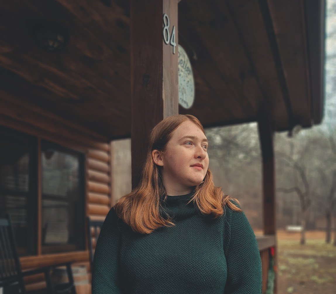 A woman with red hair and a black sweater standing outside near a wooden building, looking to the right.