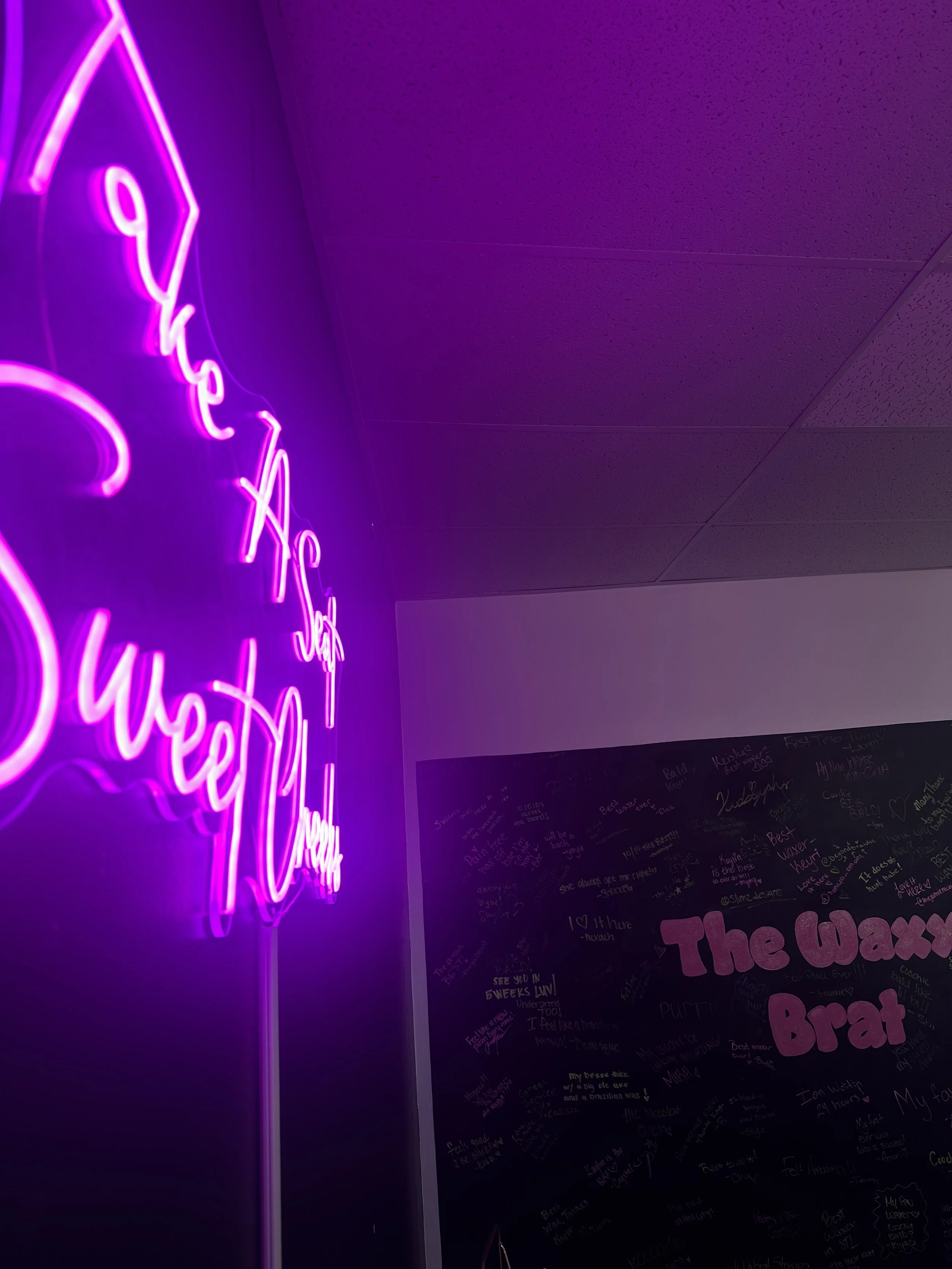 Neon pink sign reading 'Take A Seat Sweet Cheeks' on a dark green wall in a dimly lit room.