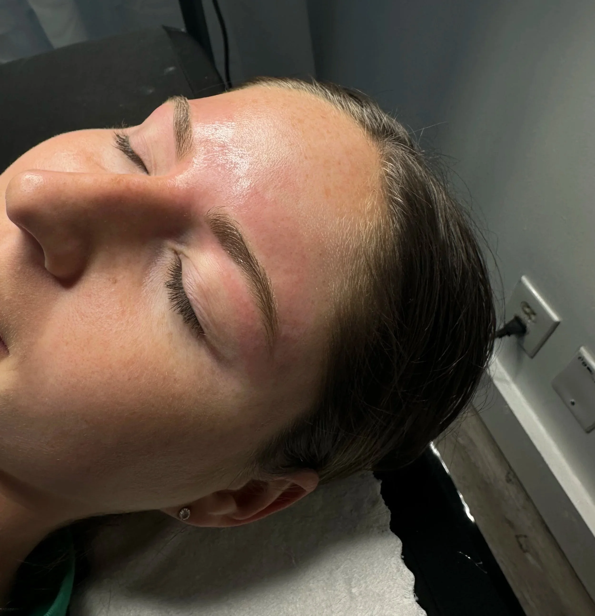 Close-up of a woman with freshly done eyebrow wax, lying on a treatment table with her eyes closed.