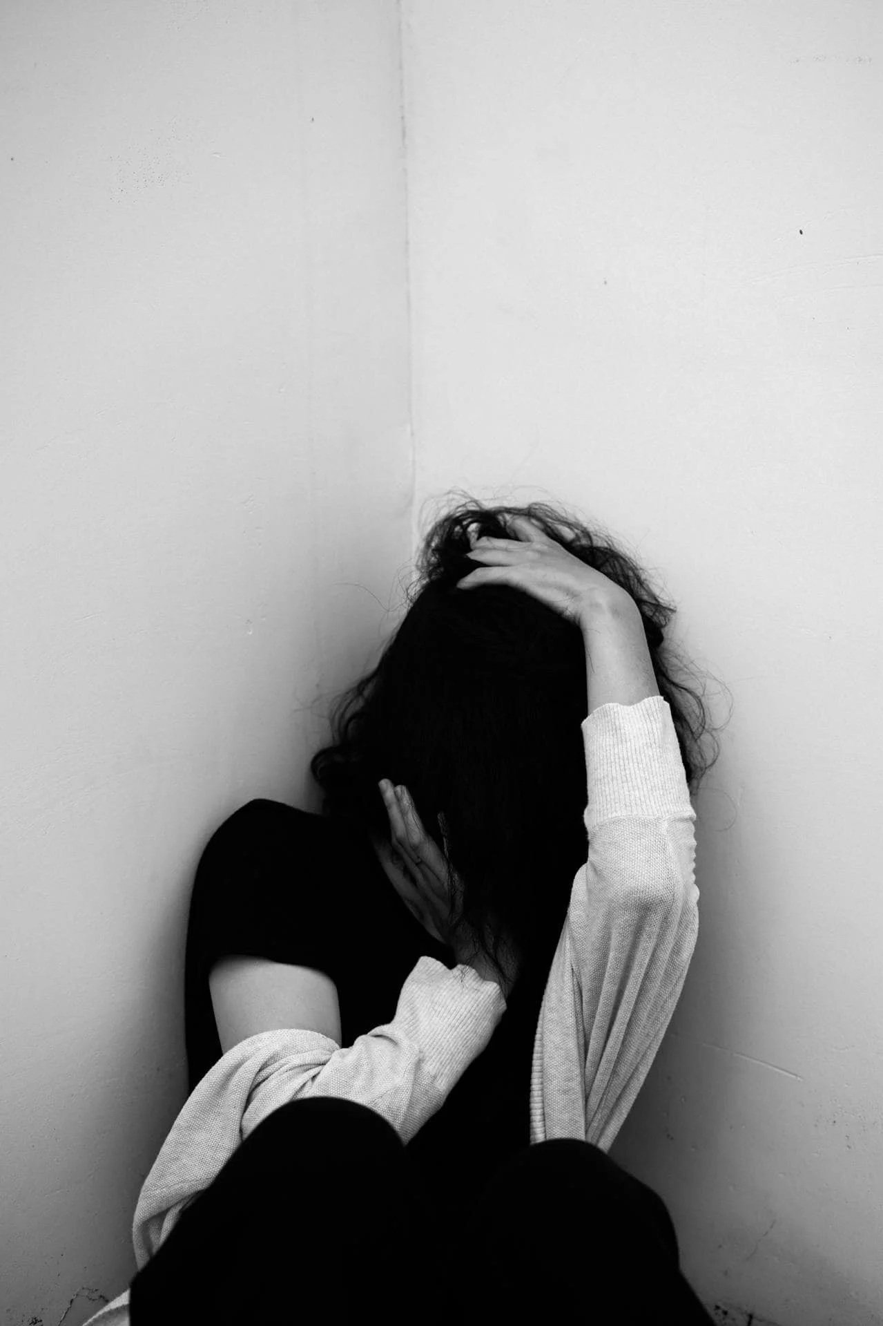 Person with dark, curly hair sitting against a plain wall, head bowed, hands in hair or holding head, wearing a black shirt and a light-colored long sleeve shirt.