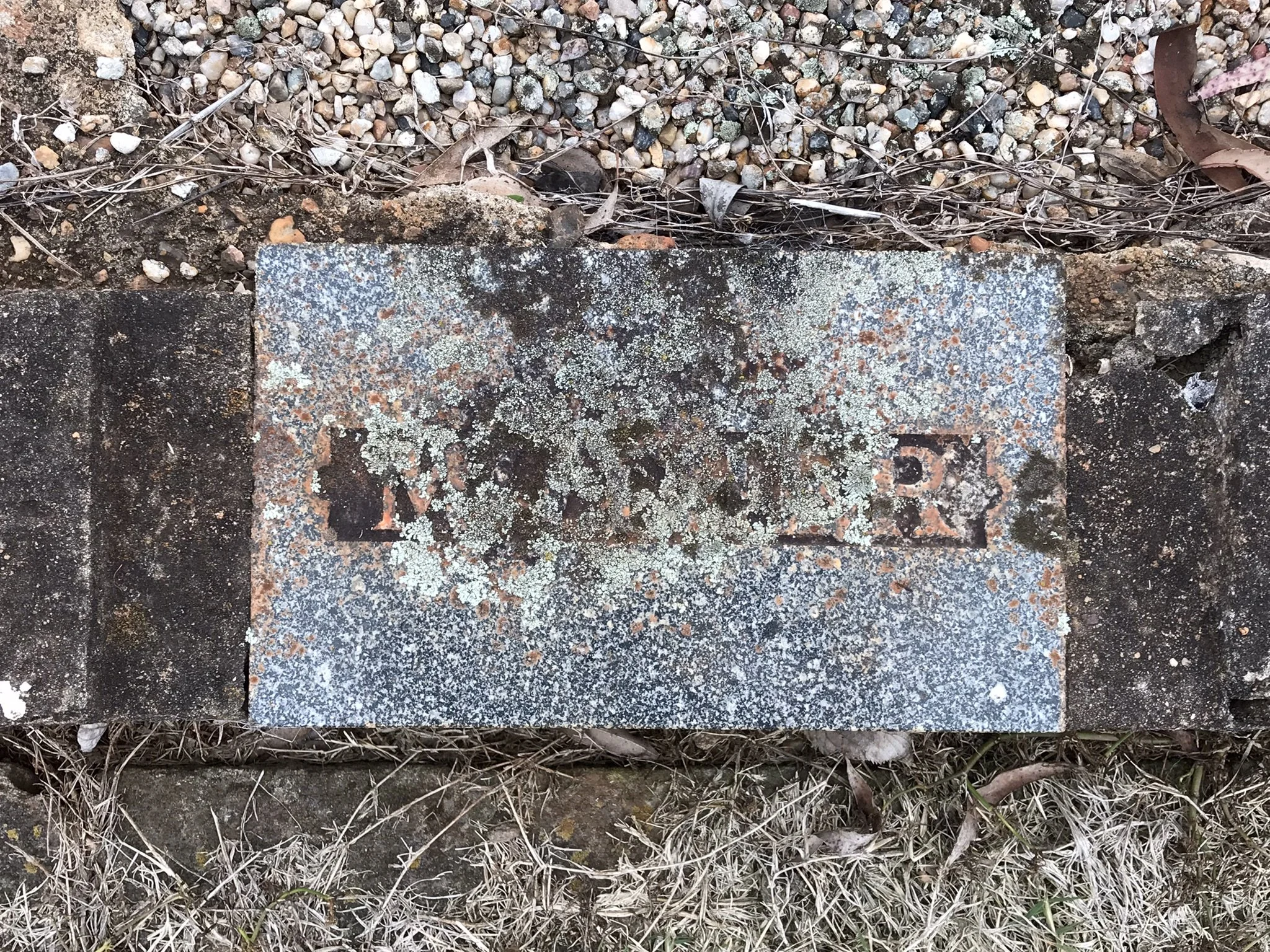 A weathered, rusty brick with the remnents of the etched word 'MOTHER' on it, surrounded by gravel, dirt, and dried grass.