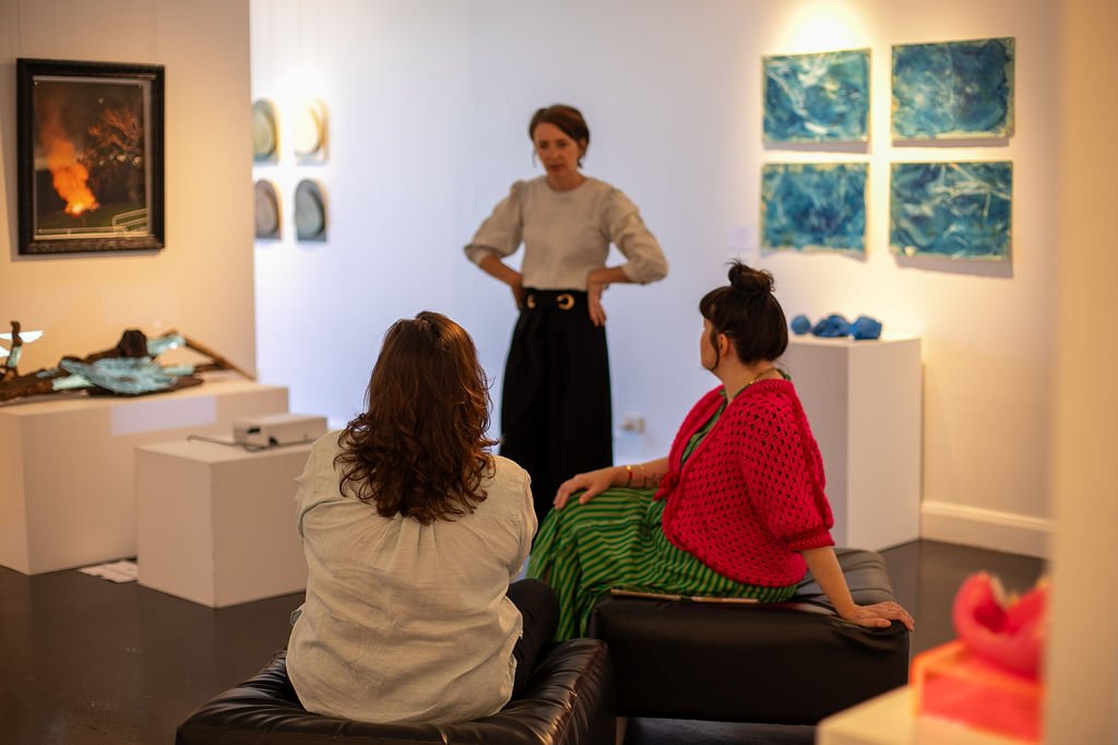 Three women are inside an art gallery with paintings on the walls. One woman stands talking, while two women sit on a black sofa; one wears a red jacket and green striped skirt, the other has a beige shirt. The gallery has white walls and dark floori