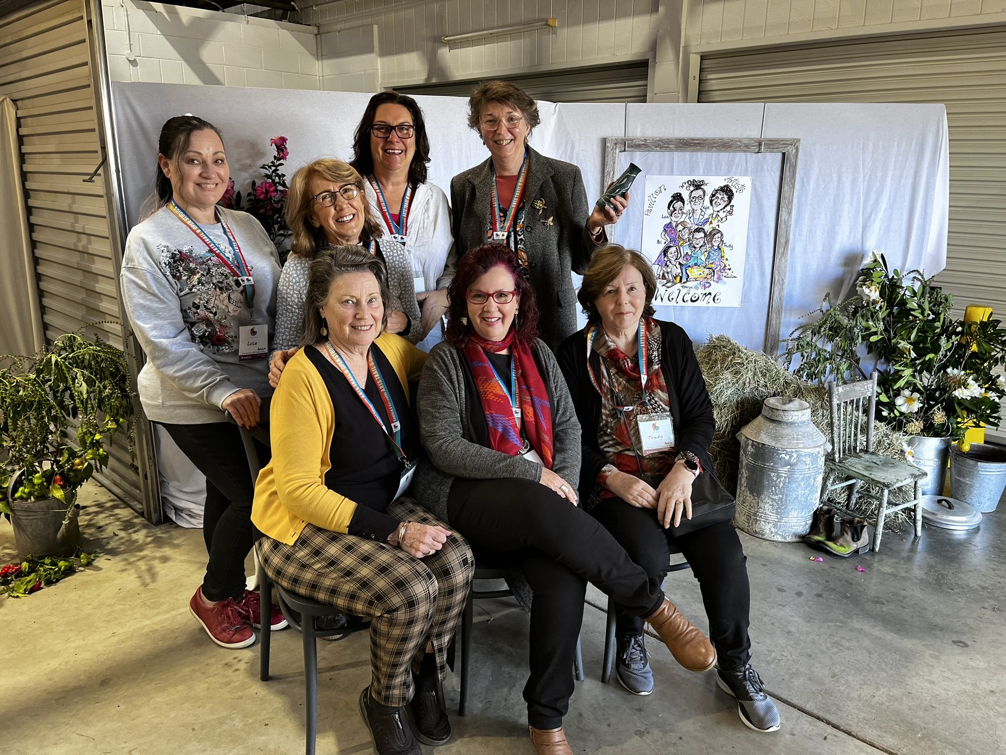 Group of nine women smiling and posing for a photo at an indoor event, with some seated and others standing. They are wearing badges and colorful clothing, and there are plants and decorative items in the background.