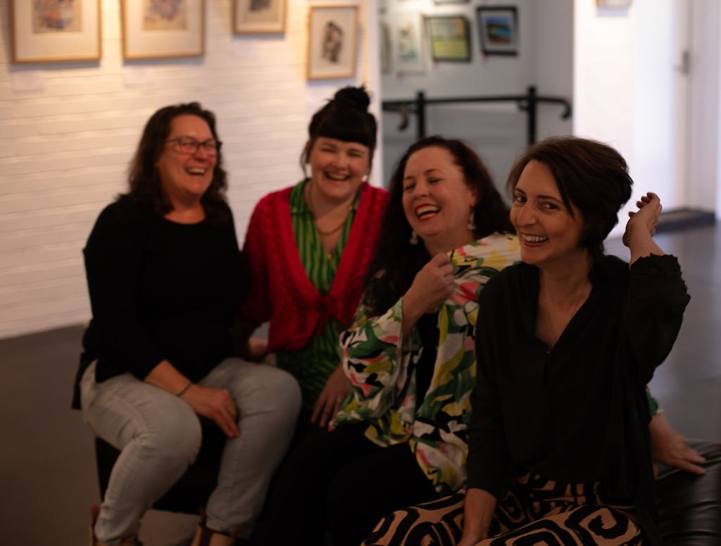 Four women sitting together, laughing and enjoying each other's company in an art gallery or exhibition space with framed pictures on the wall behind them.