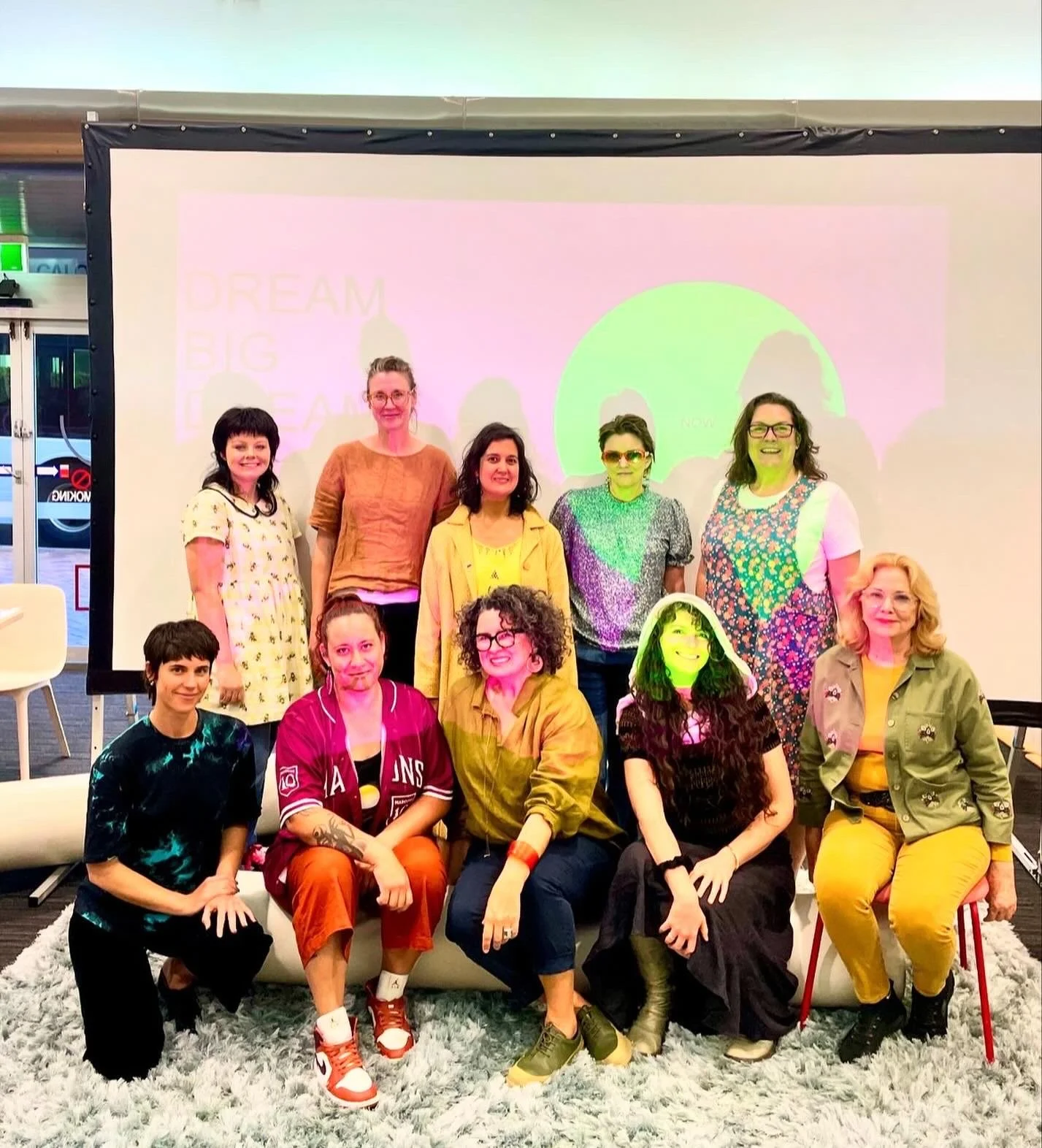 A group of ten women posing together indoors in front of a large screen, smiling and dressed in colorful outfits, with a pink and green slide visible on the screen behind them.