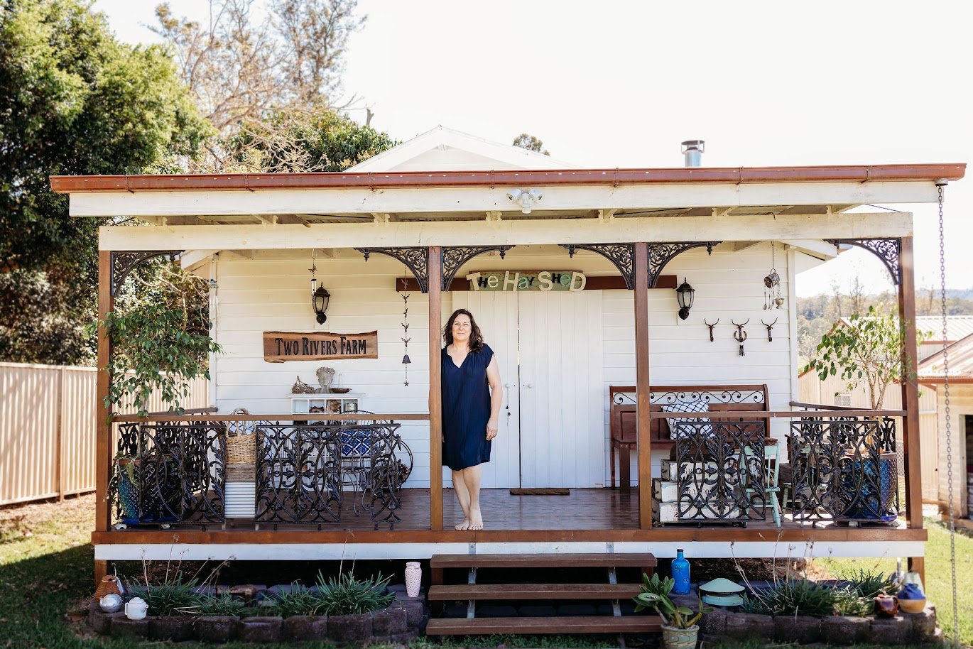A woman in a navy blue dress standing on the porch of a white house with decorative black metal railings. The house is decorated with signs, lanterns, and antlers, and has a porch swing and outdoor furniture. There are trees and a wooden fence in the