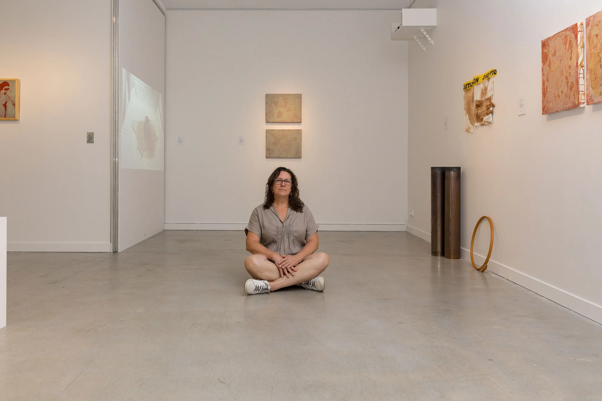 A woman sitting cross-legged on the floor of an art gallery, with abstract paintings and art pieces on the white walls behind her.