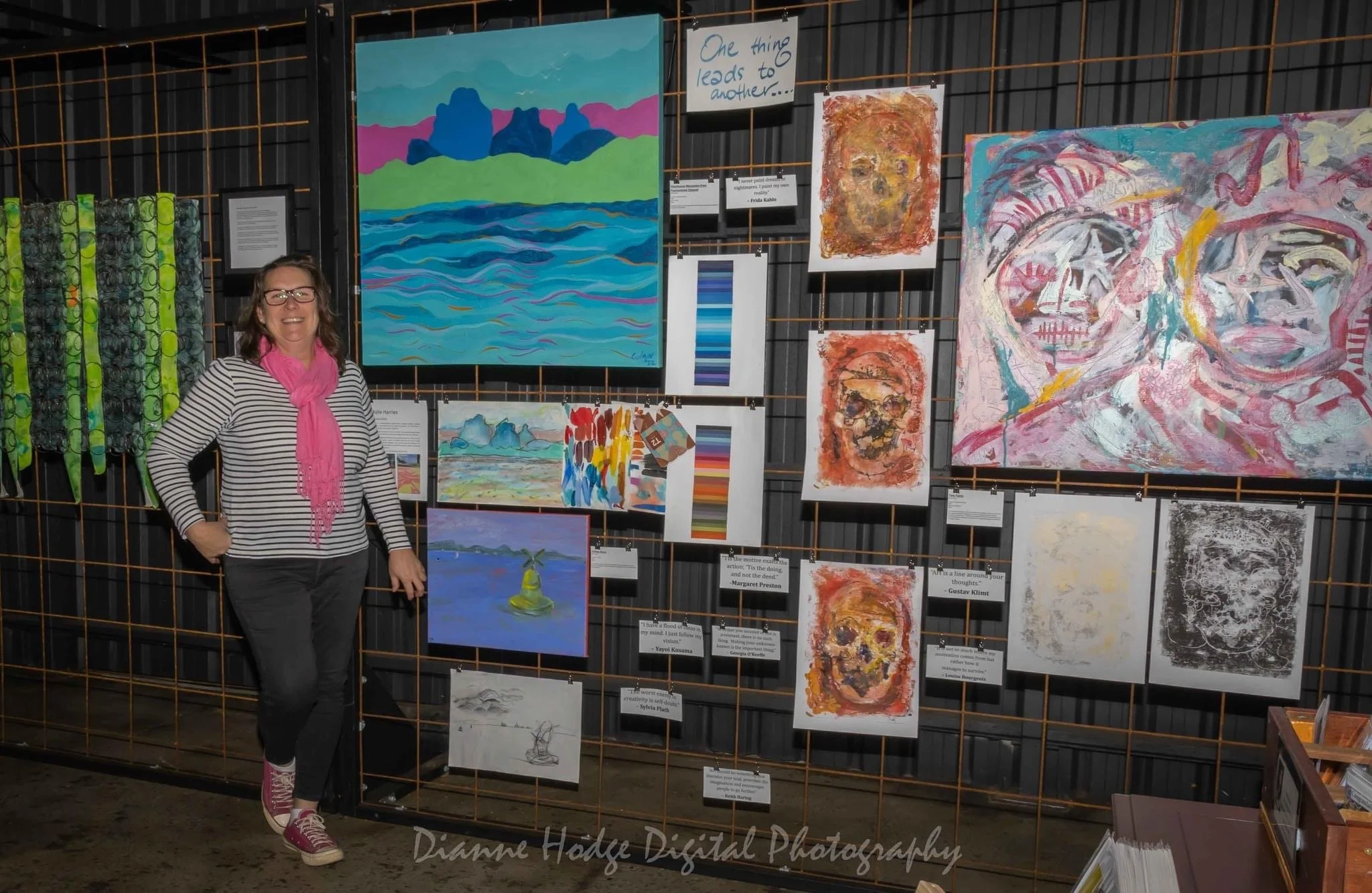 A woman smiling and dressed in a striped top, pink scarf, and gray pants, standing beside a display of colorful artwork on a wire grid at an art exhibit. The artwork includes abstract and landscape paintings, along with quotes and color samples.