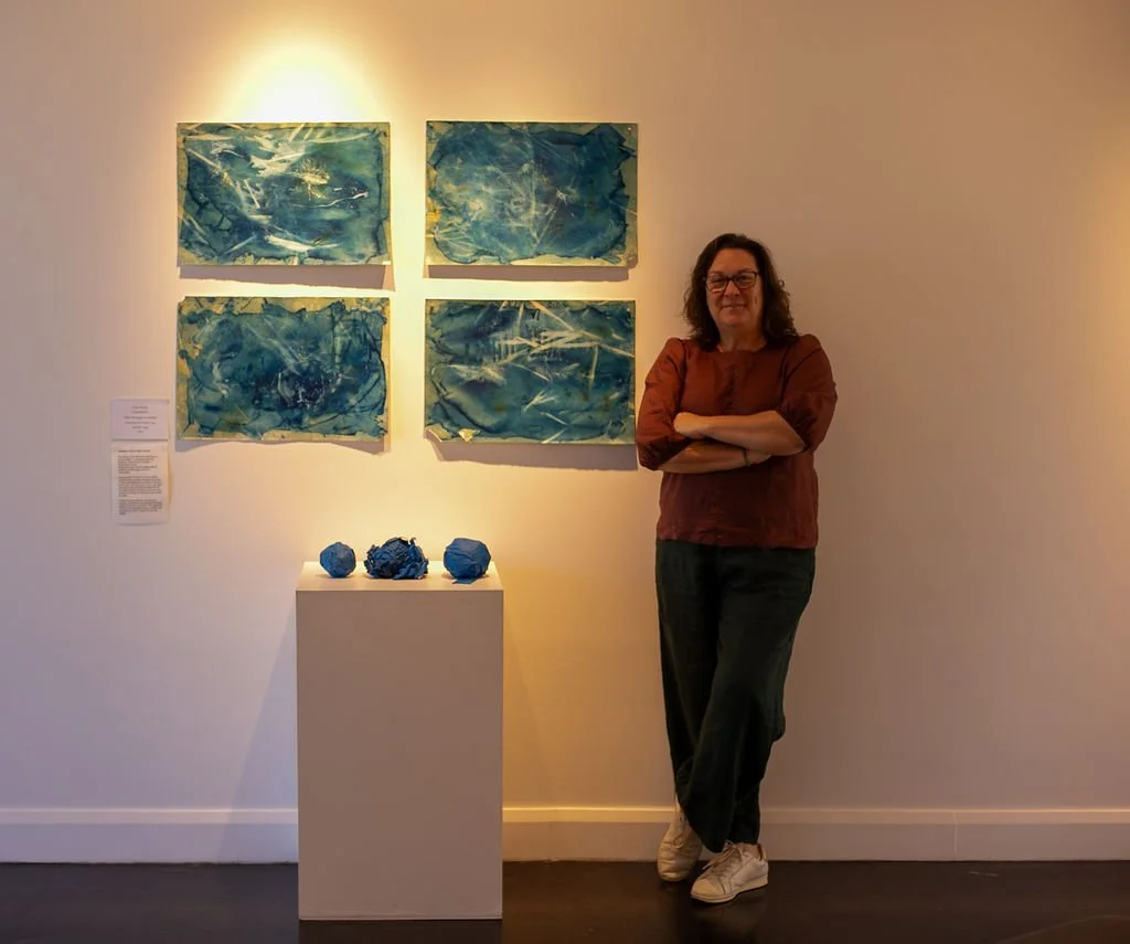 A woman stands with arms crossed in front of artwork featuring four abstract blue paintings on a white gallery wall. A small white pedestal holds three crumpled blue paper sculptures.