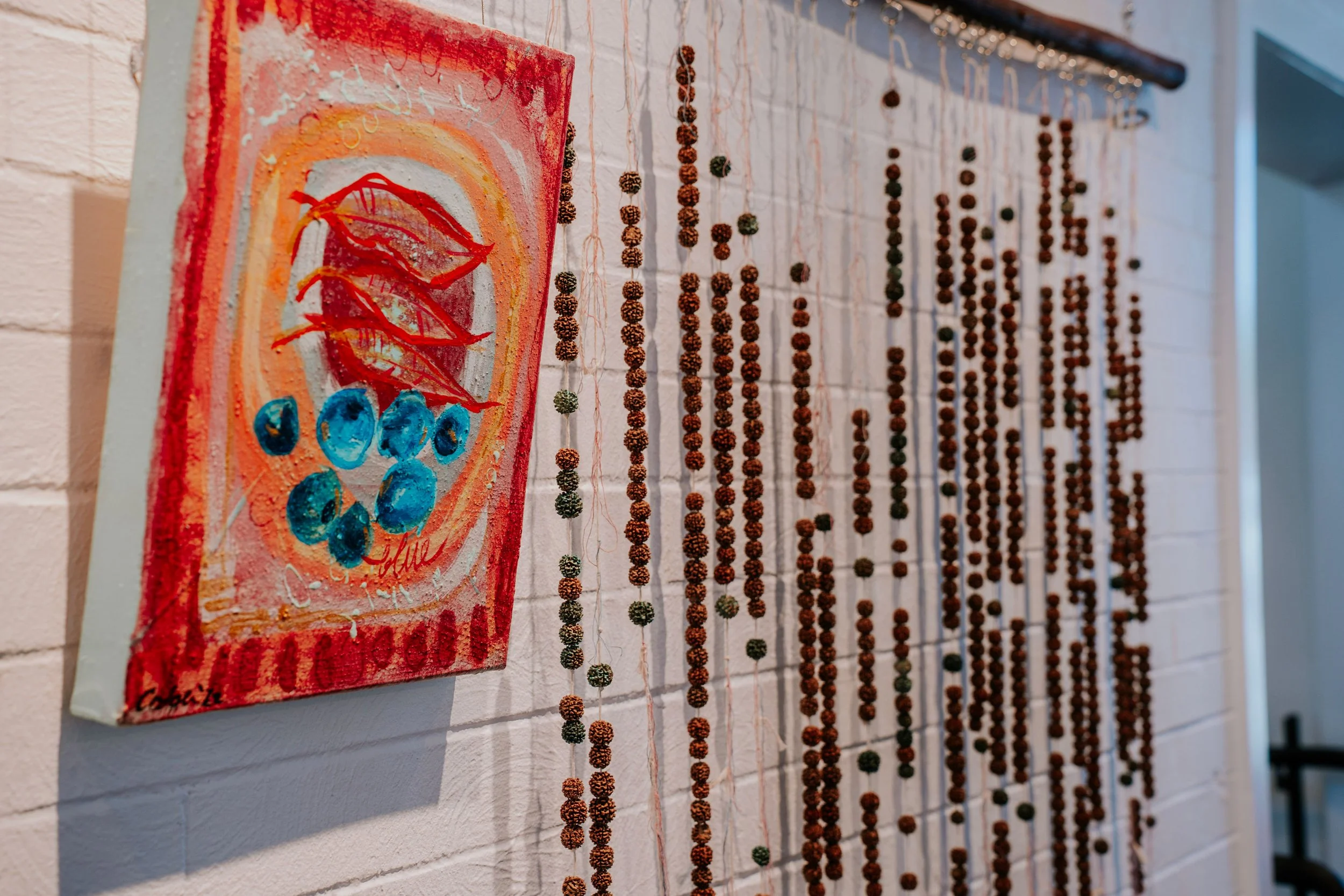 A small abstract colorful painting on a white brick wall, featuring red lips, blue circles, and concentric circles. To the right, hanging strands of small beads in brown and green shades are displayed on hooks.